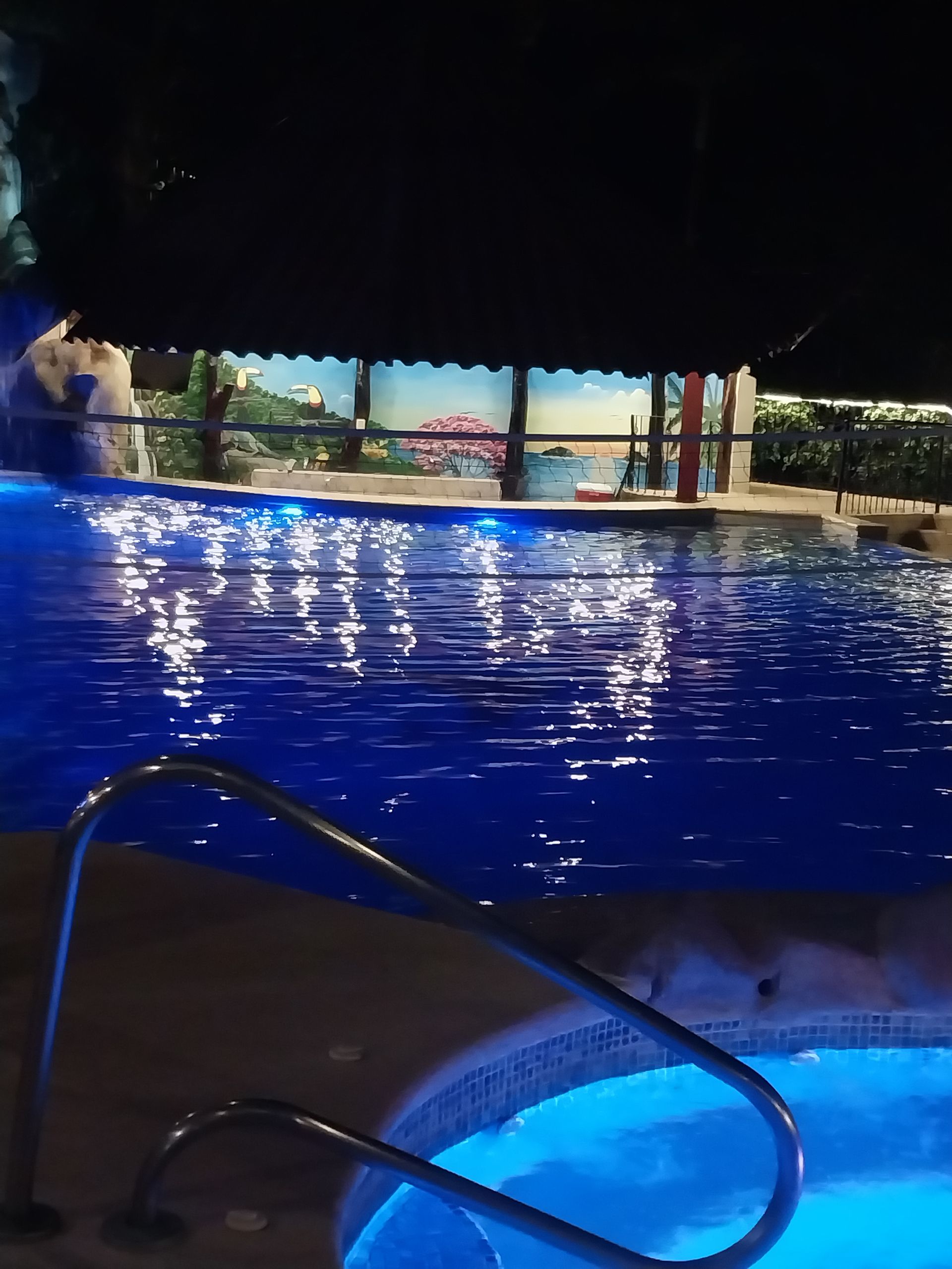 A nighttime view of a pool with blue underwater lights, a jacuzzi, and a covered patio area in the background.