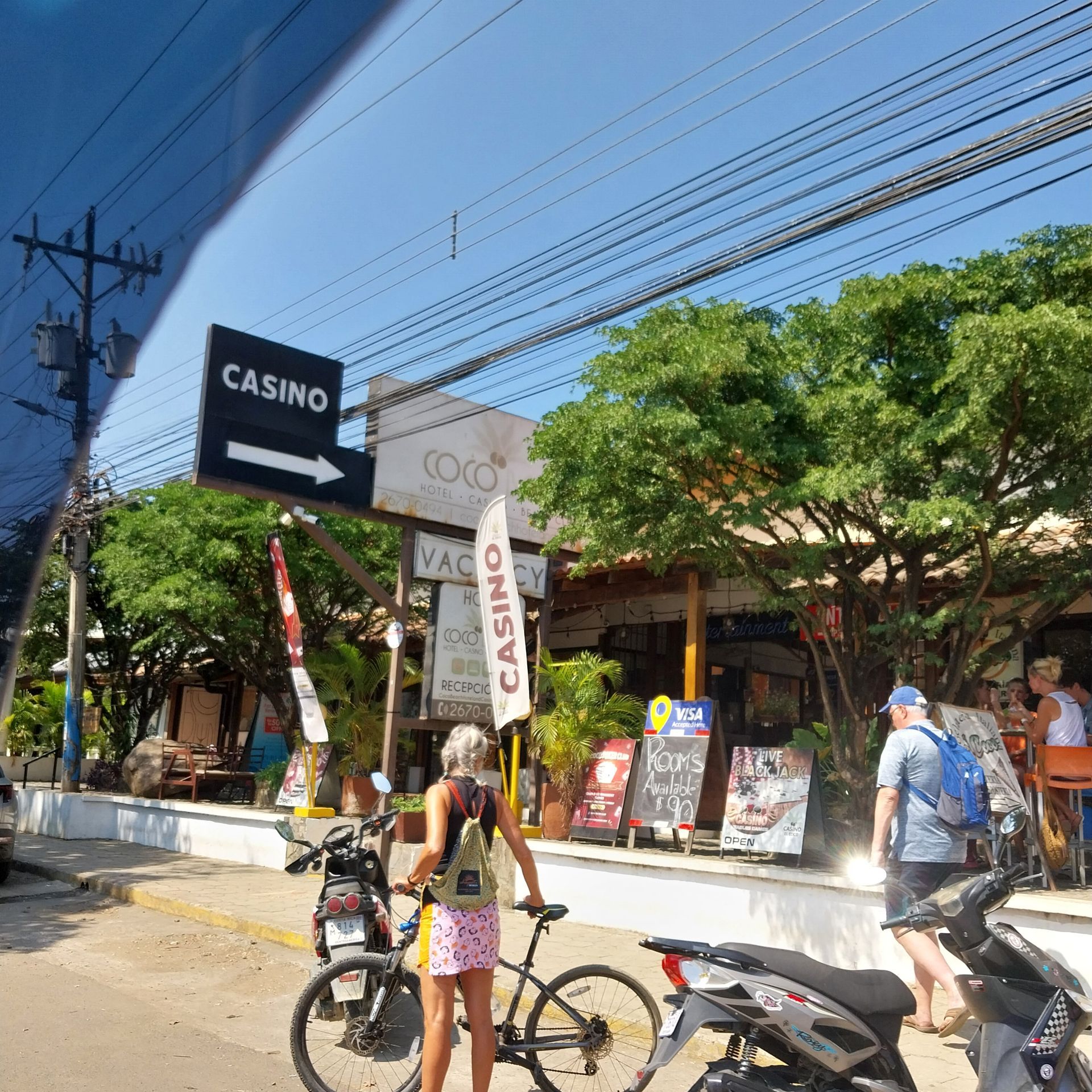 Street scene with a casino sign, people, bicycles, and storefronts on a sunny day.