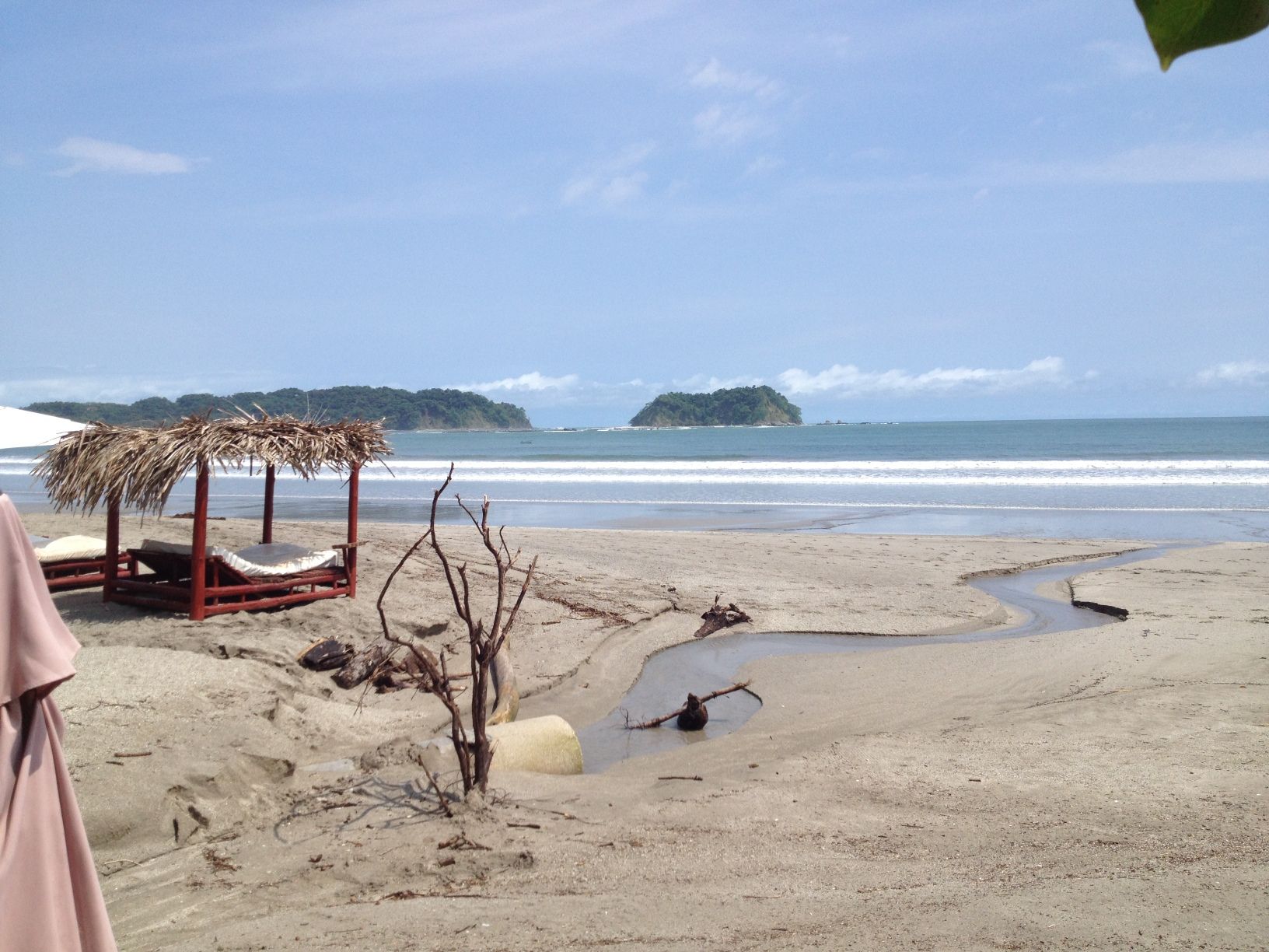 Beach scene with hut, small stream flowing to the ocean, blue sky, and islands in the distance.