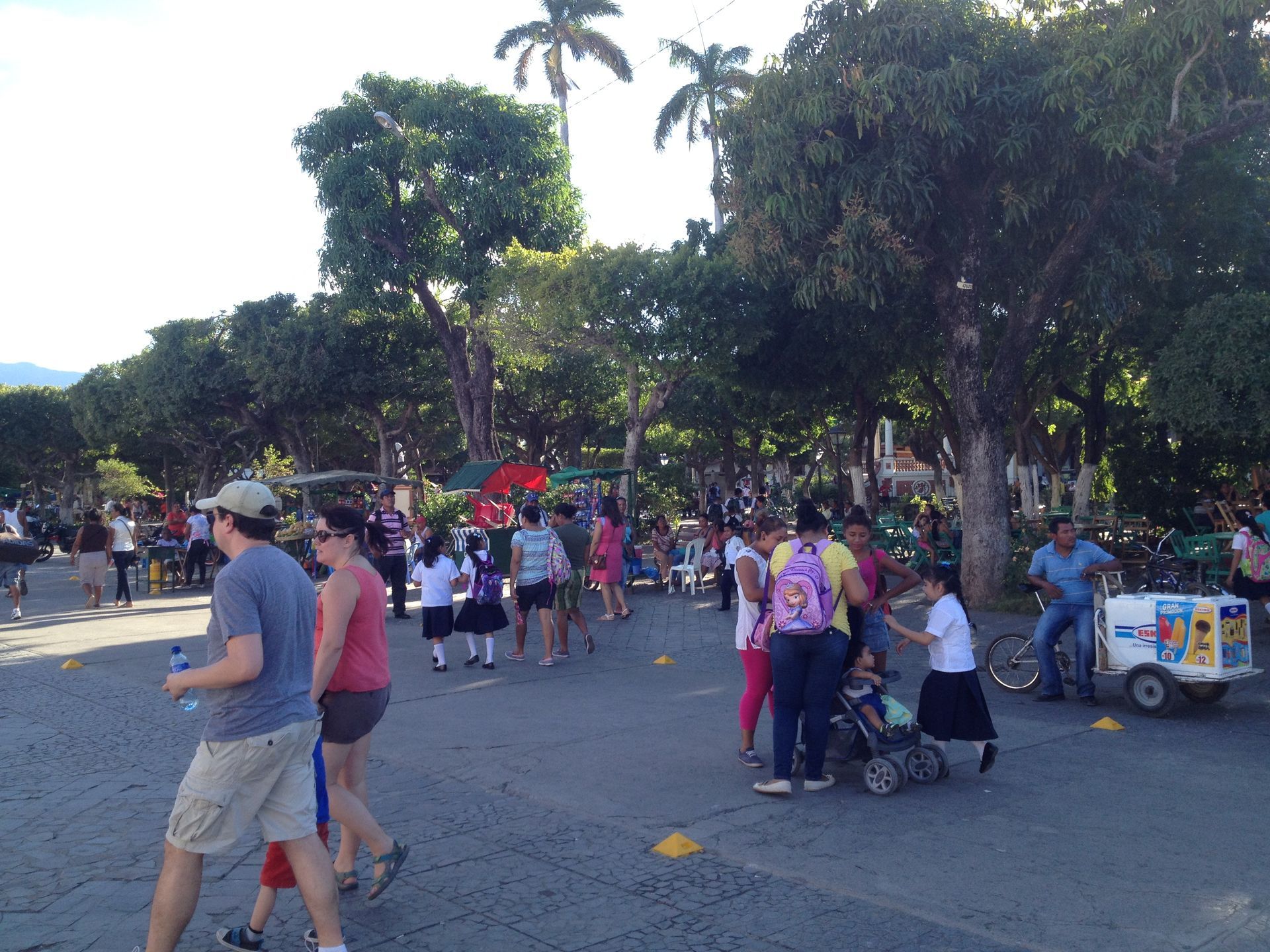 People gather in a park, possibly a community event. Ice cream cart visible. Sunny day.