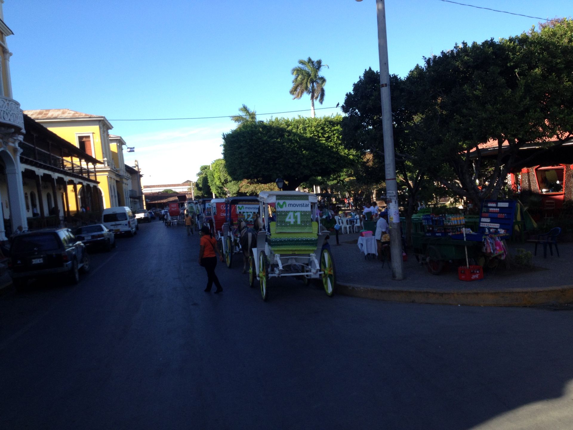 Street scene with horse-drawn carriage, people, and buildings. Green carriage, sunlight, blue sky.