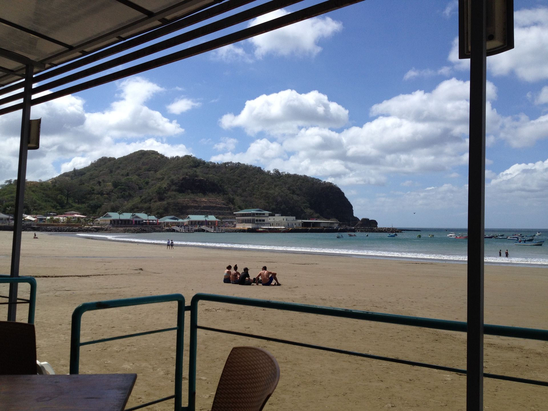 Beach scene with sandy shore, ocean, mountain backdrop, and cloudy sky. People sit on the beach; a table and railing in foreground.