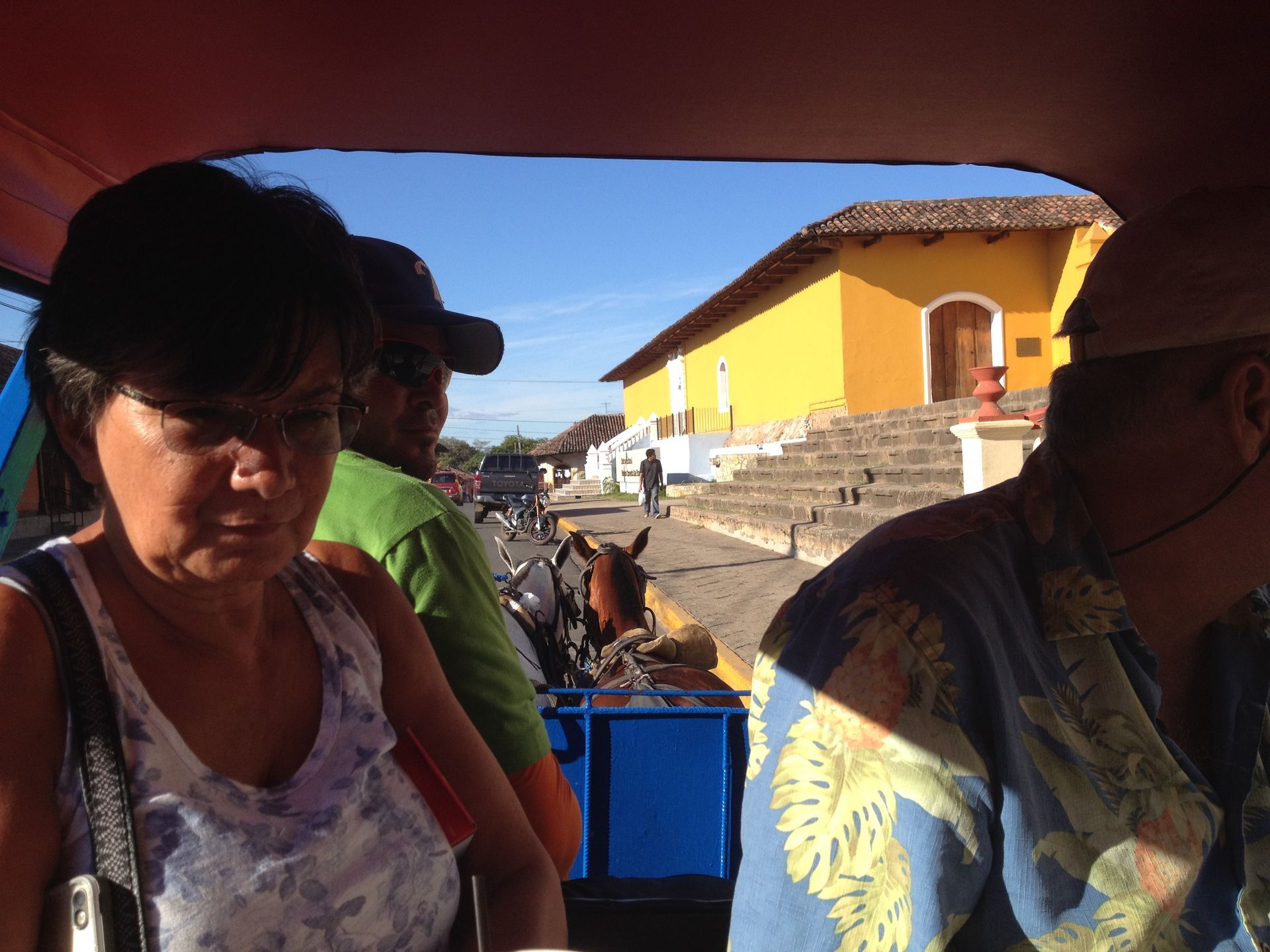 People ride in a horse-drawn carriage in front of a yellow building with steps; sunny day.