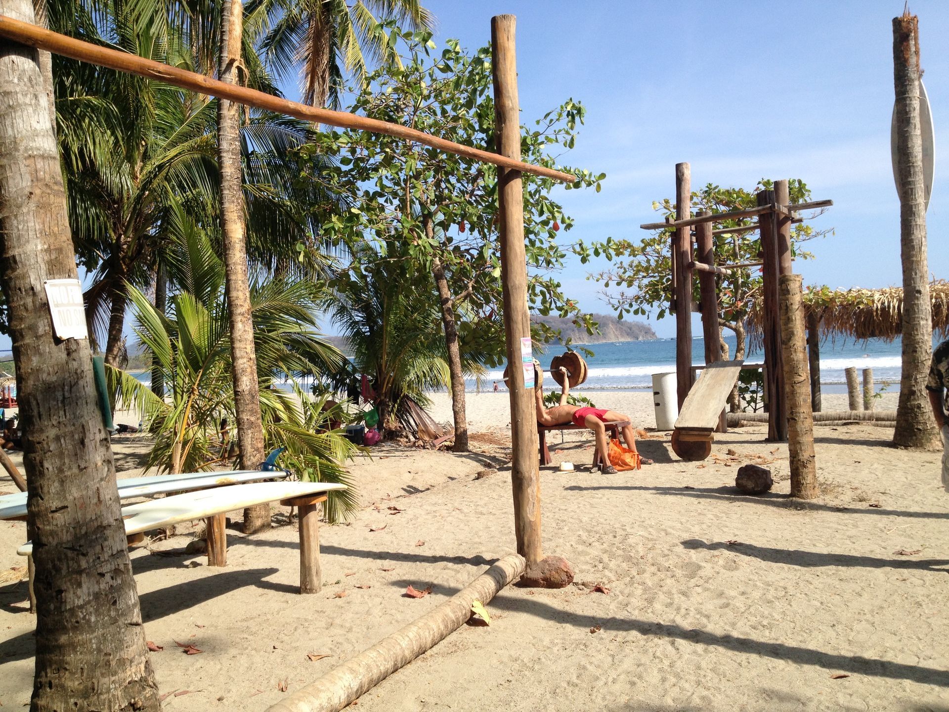 Beachside workout area: wooden pull-up bars, bench, and curved ab bench on sand; palm trees in background, sunny.