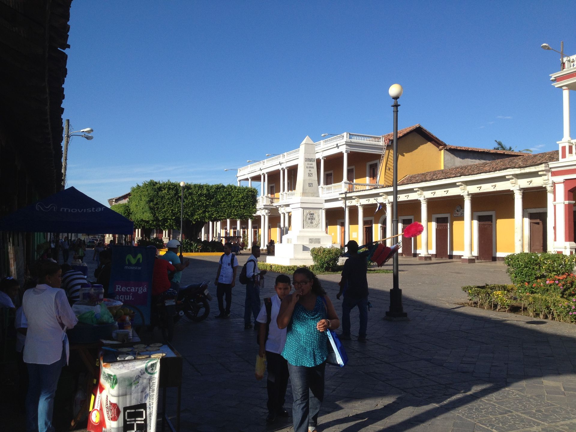Plaza with people, monument, and yellow colonial buildings under a clear blue sky.