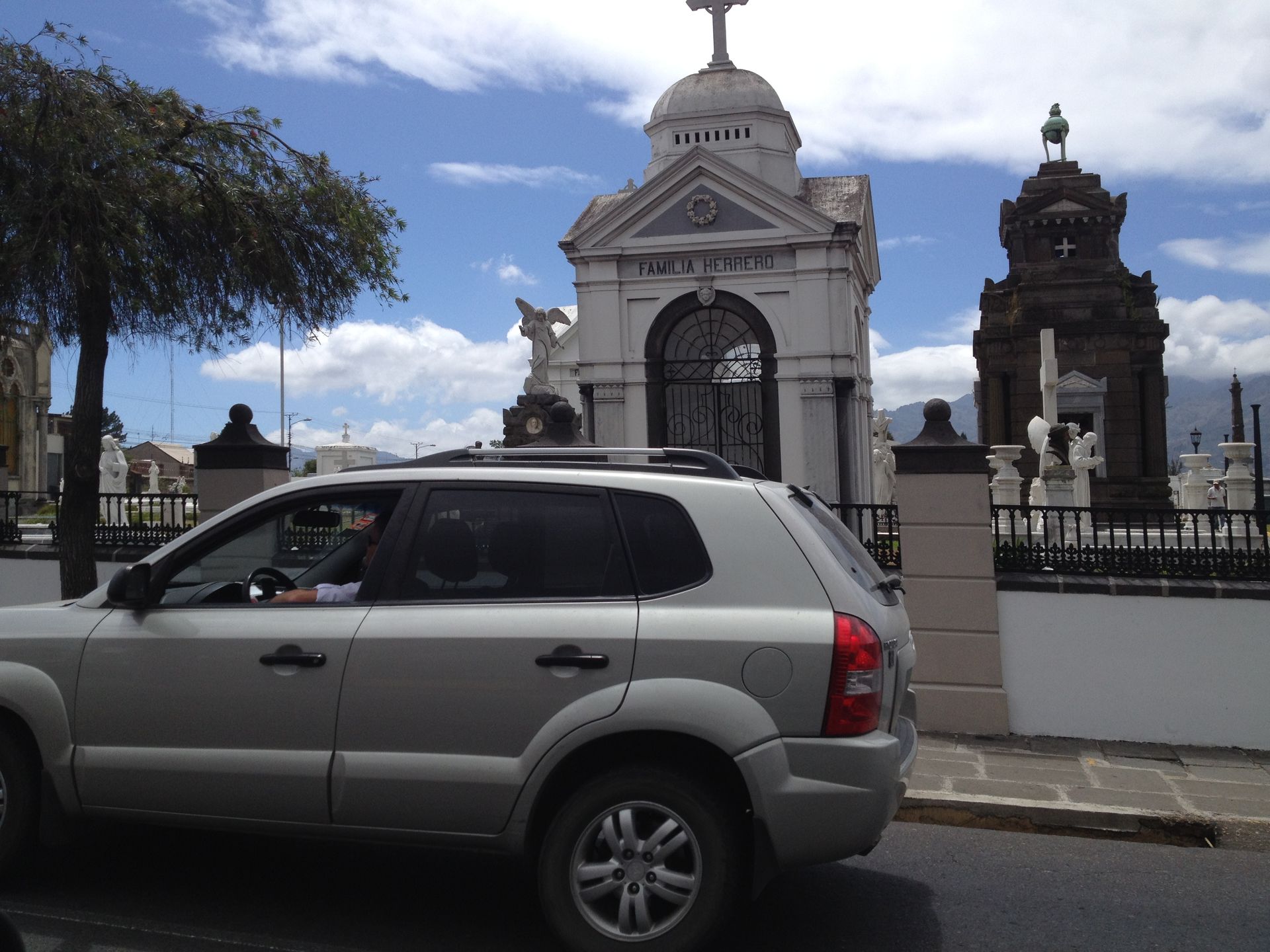 Silver SUV drives past ornate cemetery buildings under a cloudy blue sky.