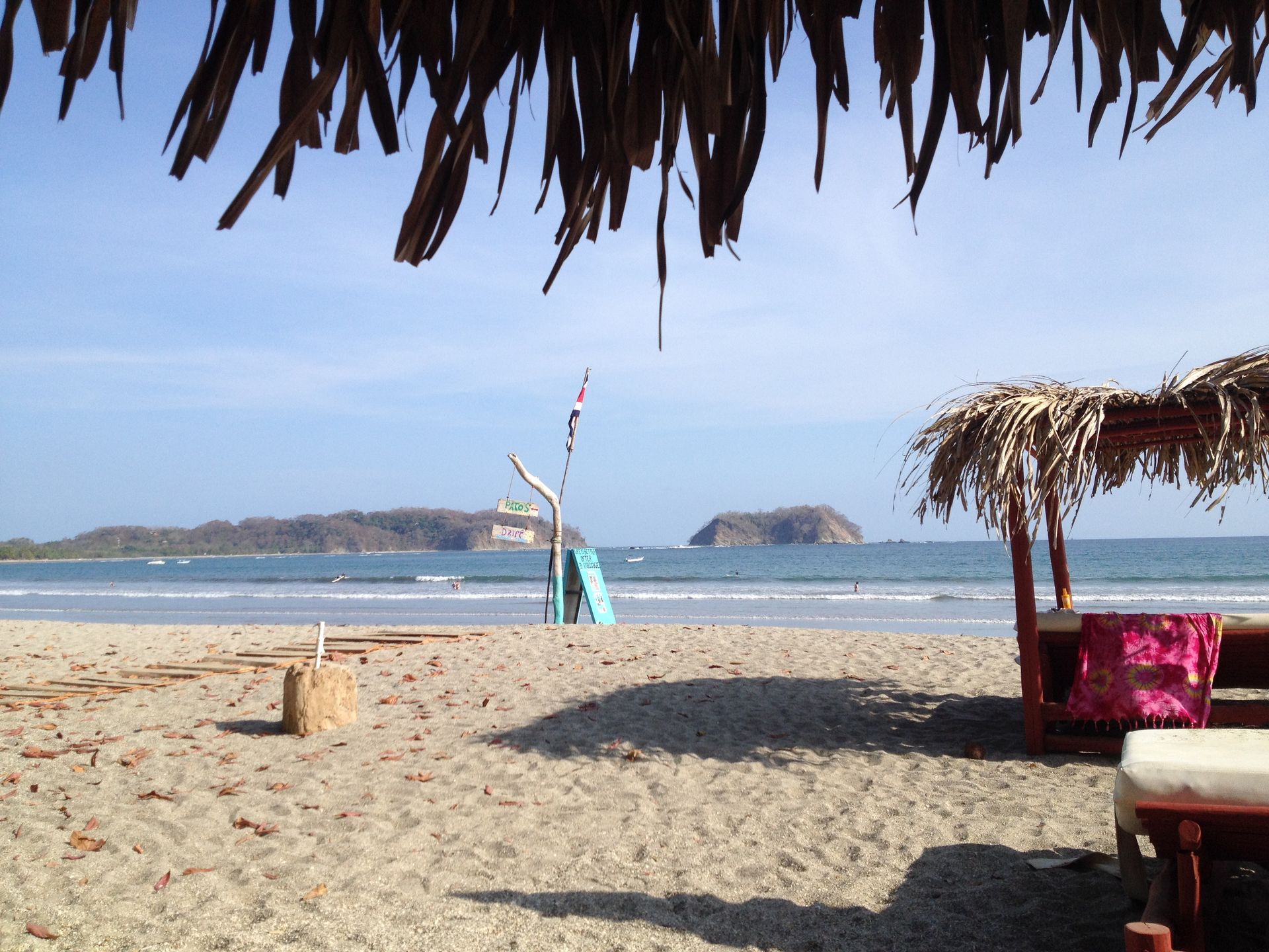 Beach scene under a thatched roof; ocean view with islands in the distance, sandy beach, lounge area.