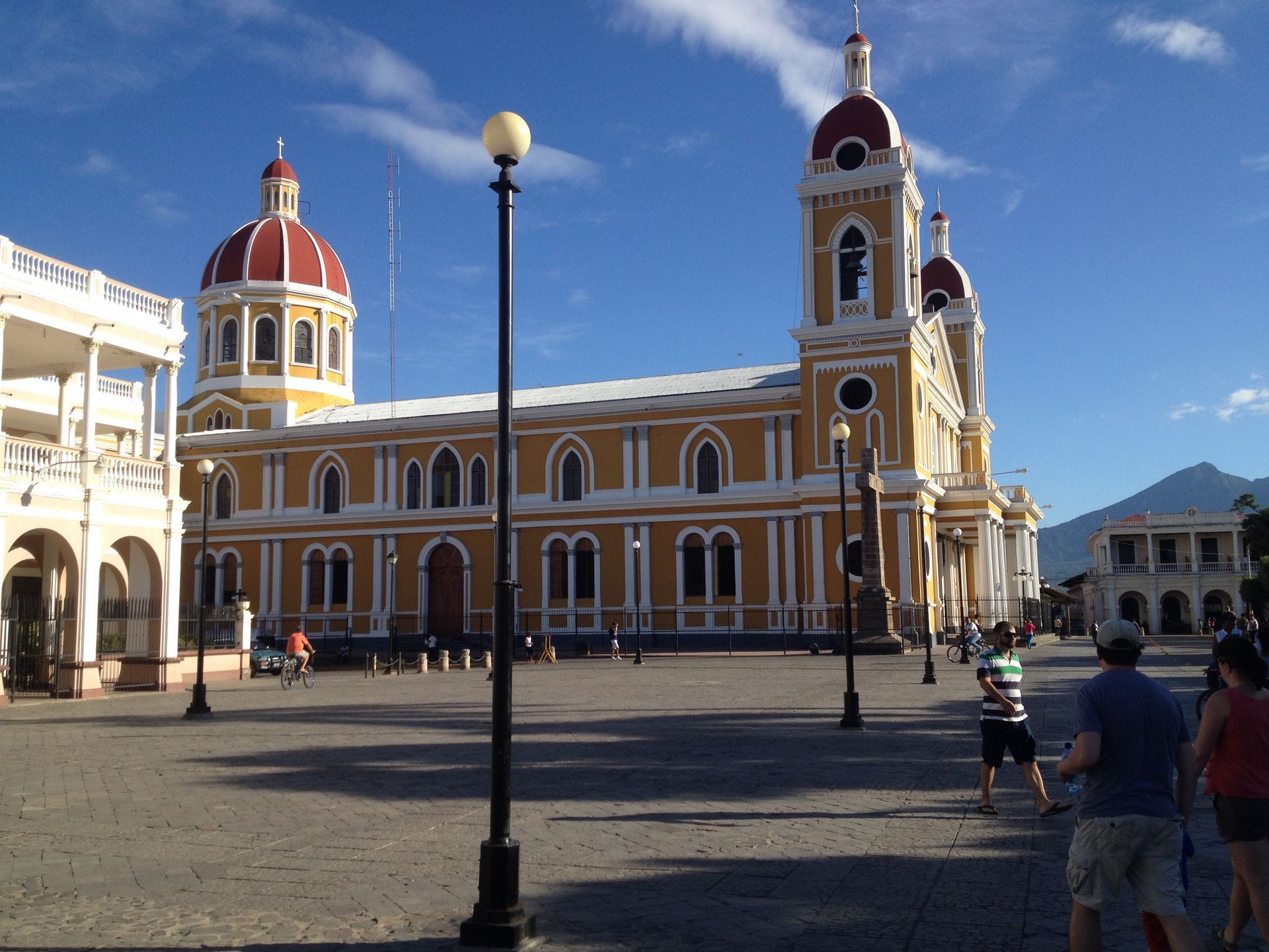 Yellow church with red domes in a plaza; people walk by on a sunny day.