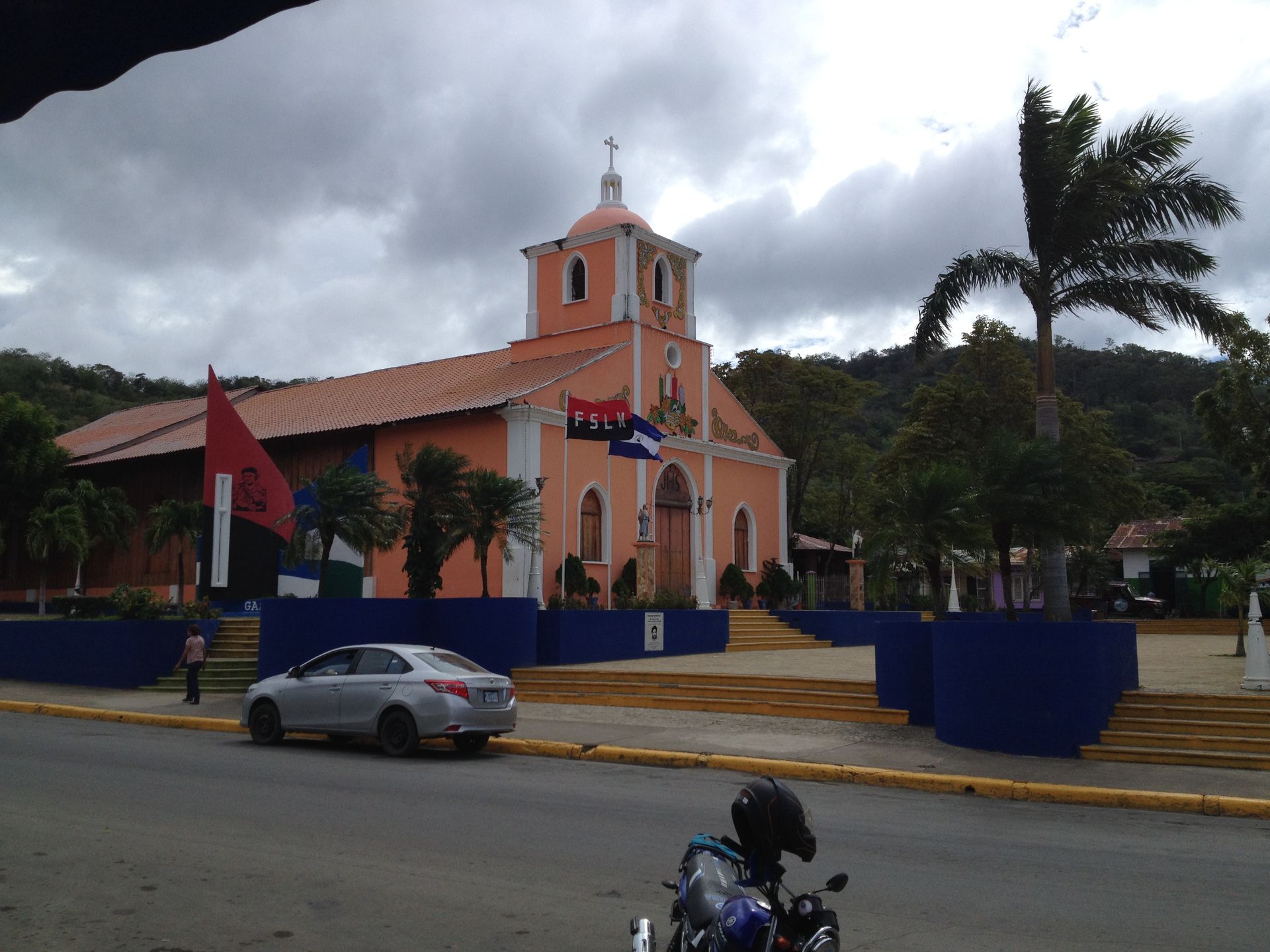 Peach-colored church with flags in a plaza, cloudy sky, car and motorcycle in foreground.