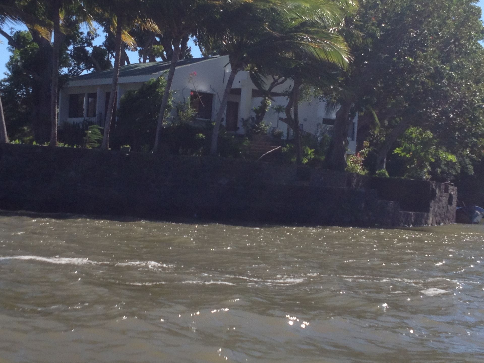 House with white walls and dark roof, behind a dark stone wall along the edge of water with small waves.