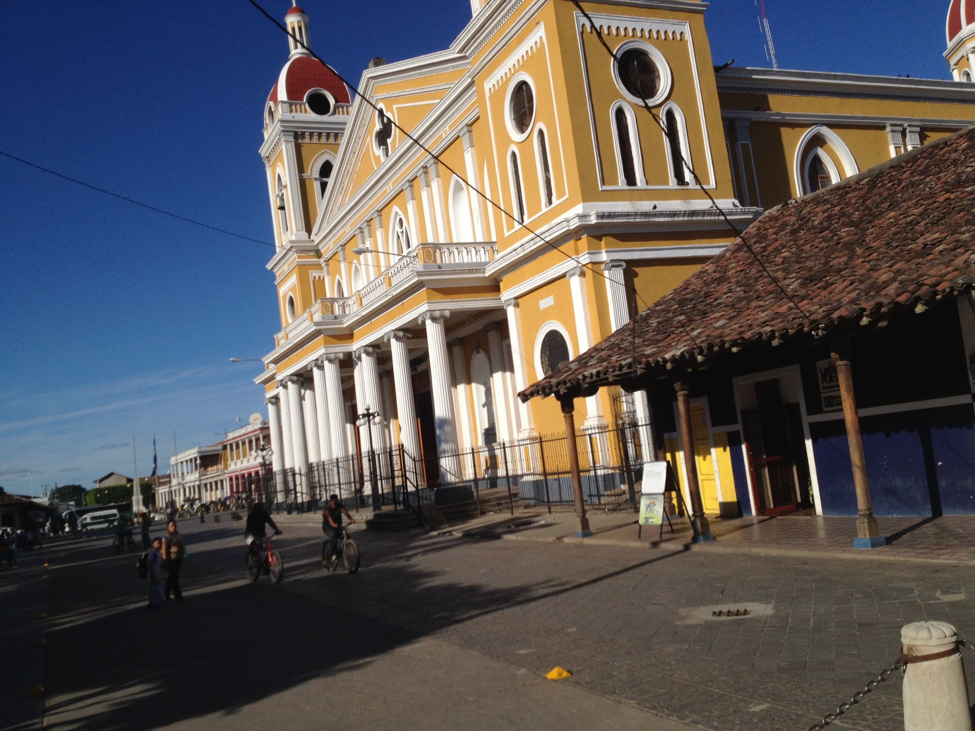 Yellow church with white columns on a city street; people biking, blue sky.