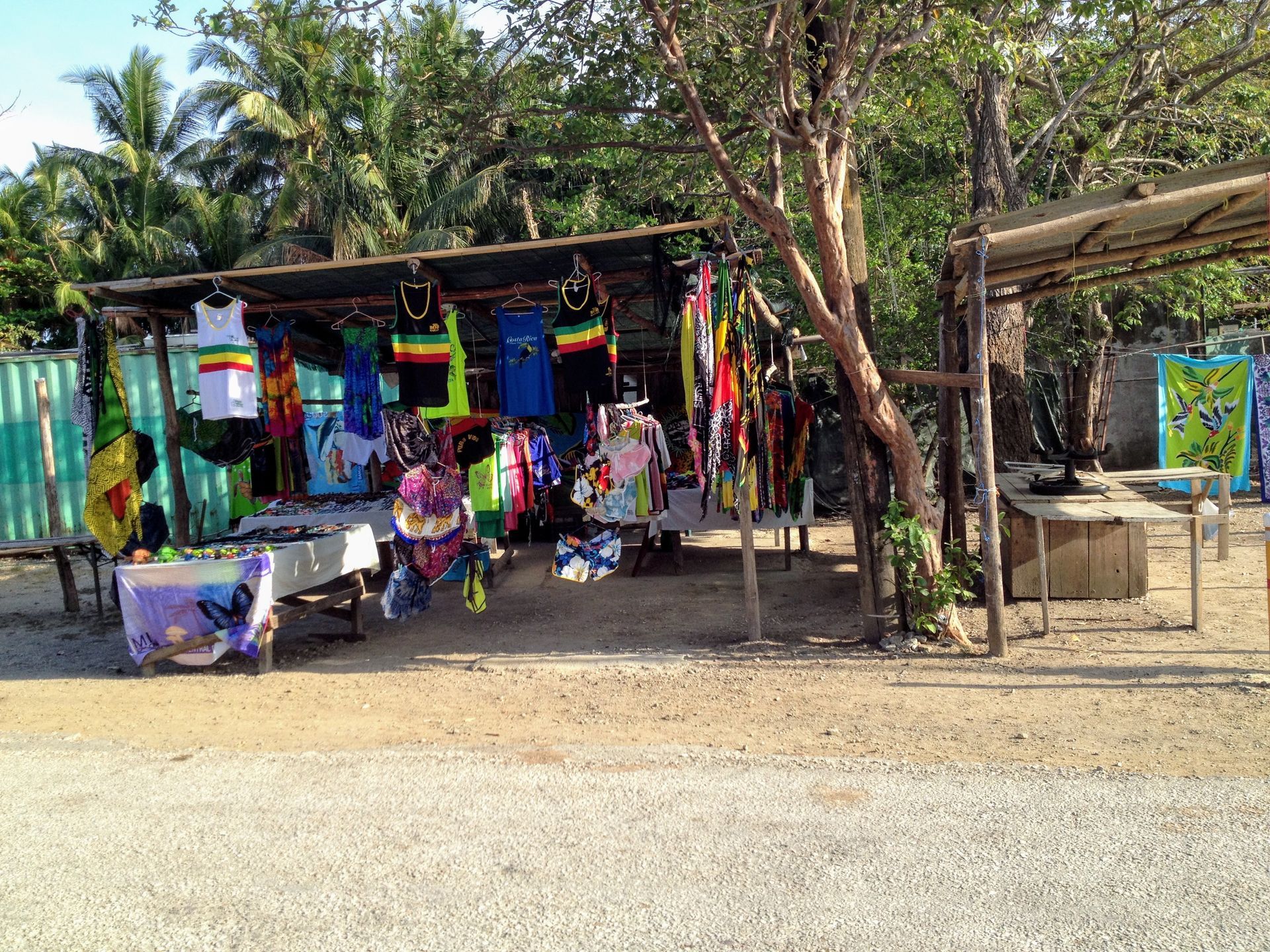 A roadside shop displays colorful clothing and souvenirs; trees and foliage in the background.