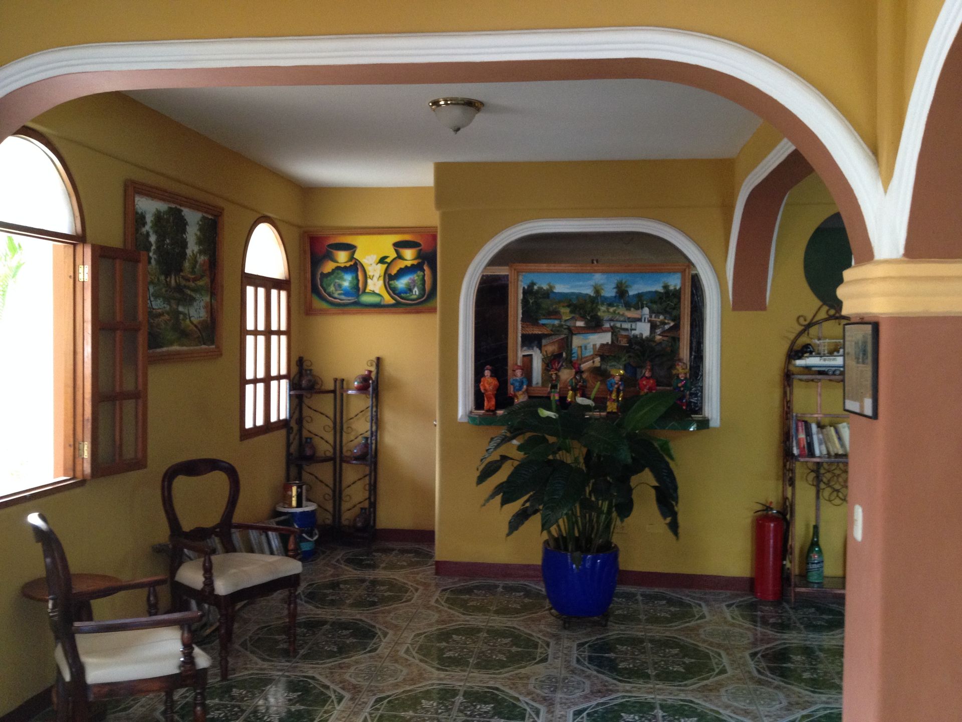 Yellow-walled room with arched doorways, art, chairs, tiled floor, and a potted plant in a blue pot.