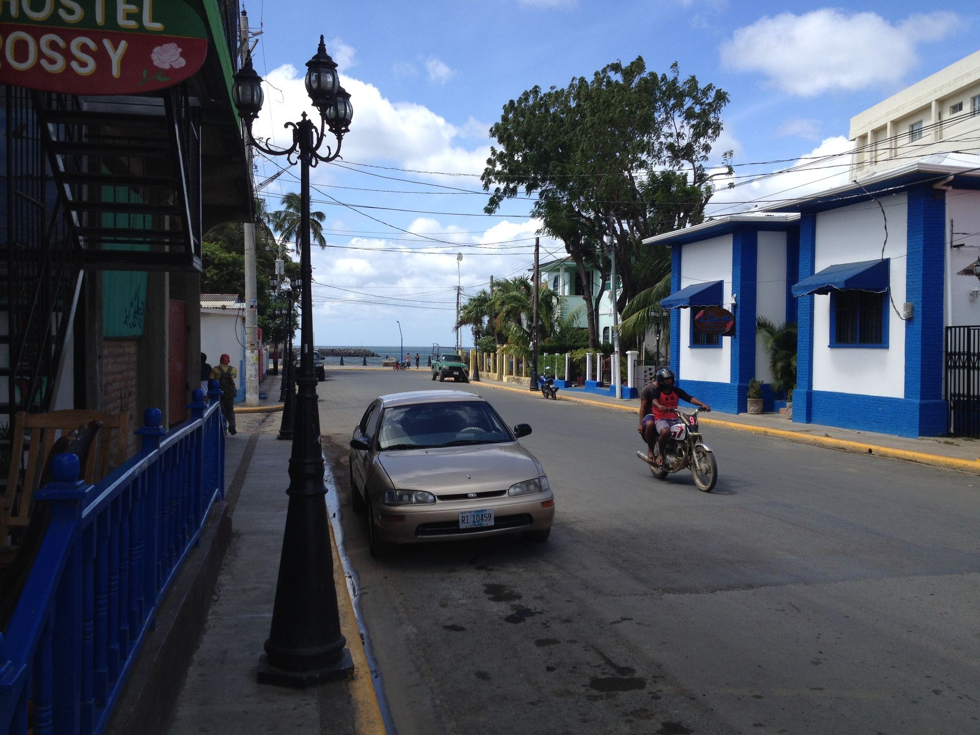 Street scene with buildings, parked car, motorcycle, and view of the sea.