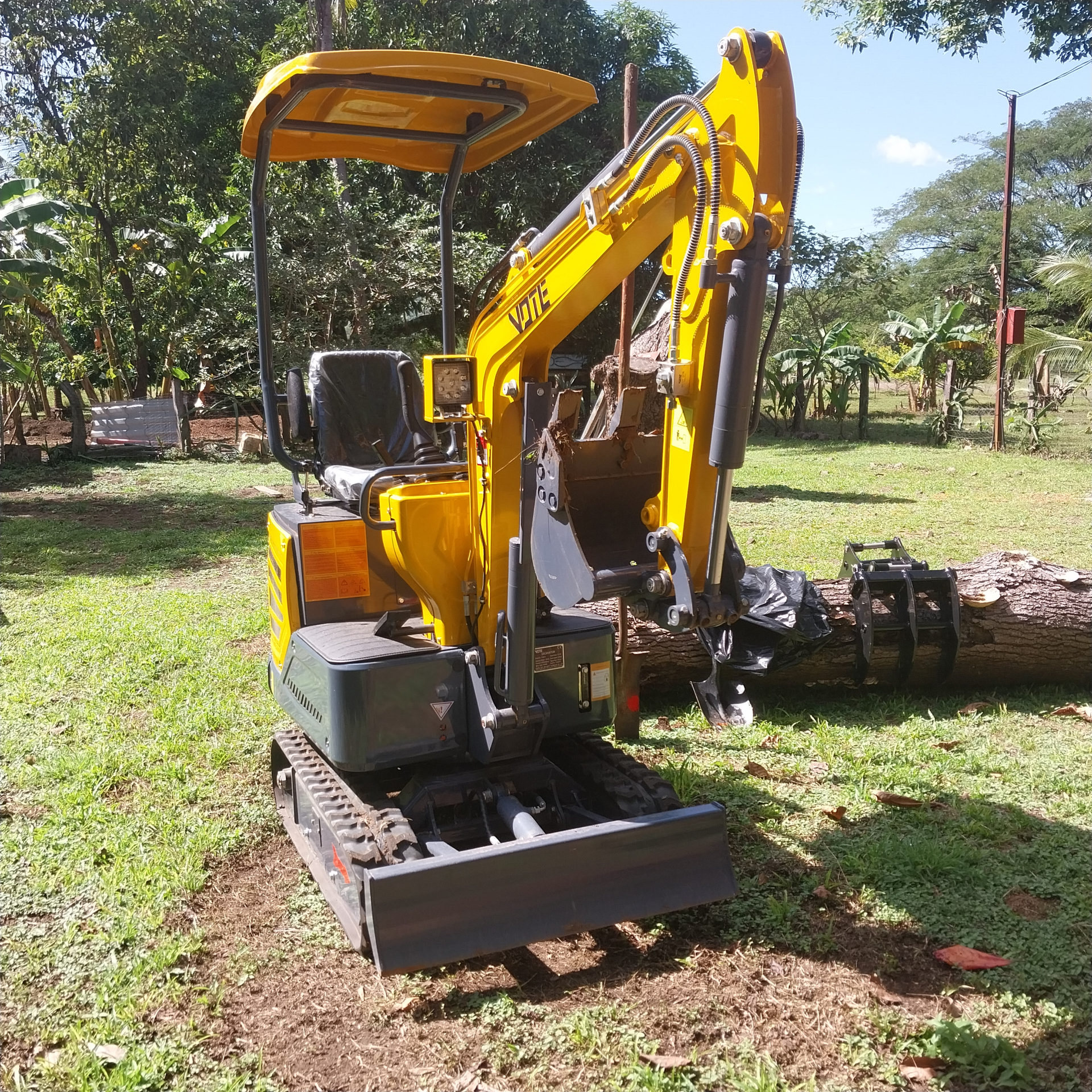Yellow mini excavator on grass, lifting a log outdoors under a sunny sky.