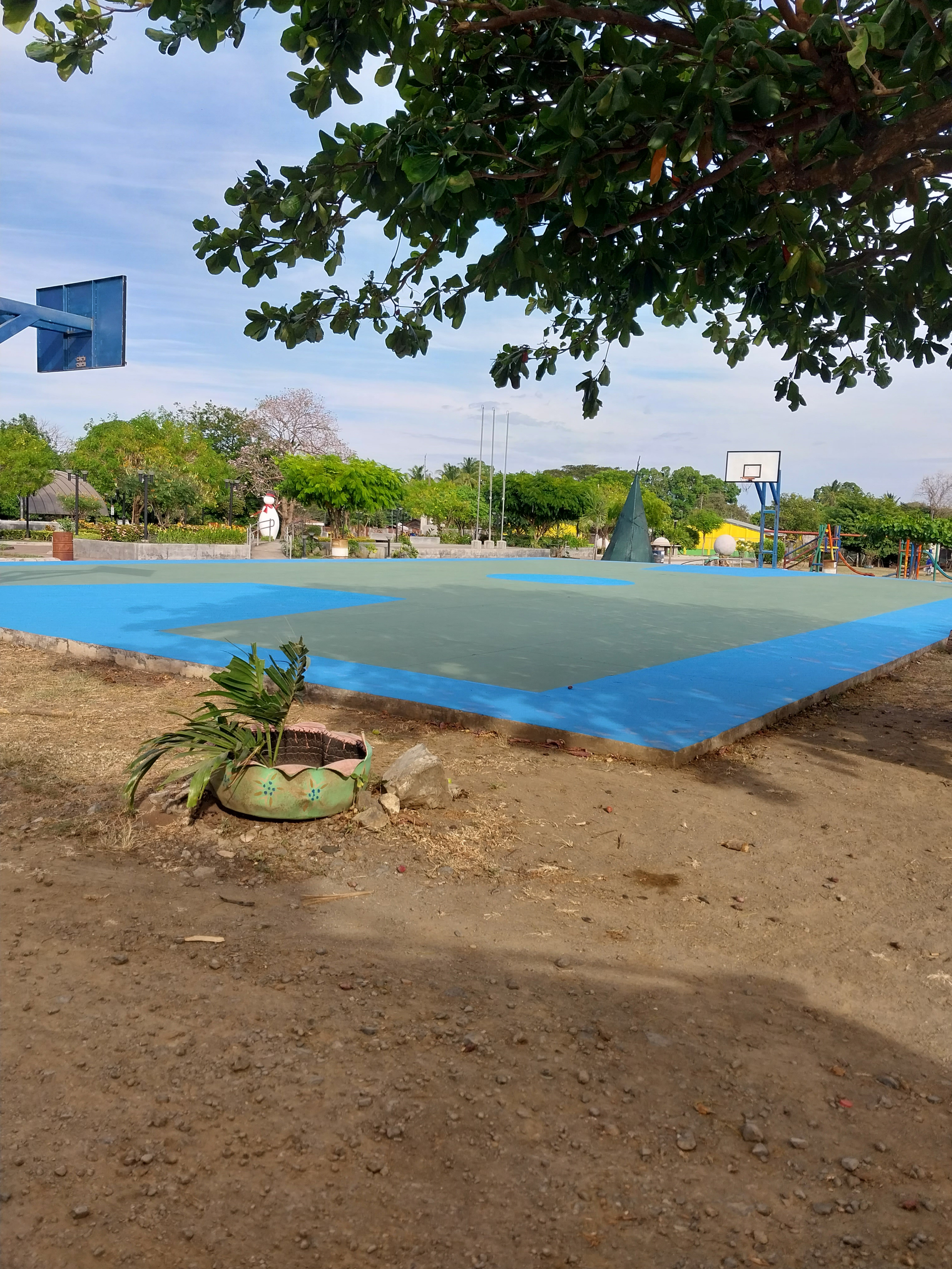 A basketball court with blue and green paint surrounded by trees and dirt ground under a blue sky.