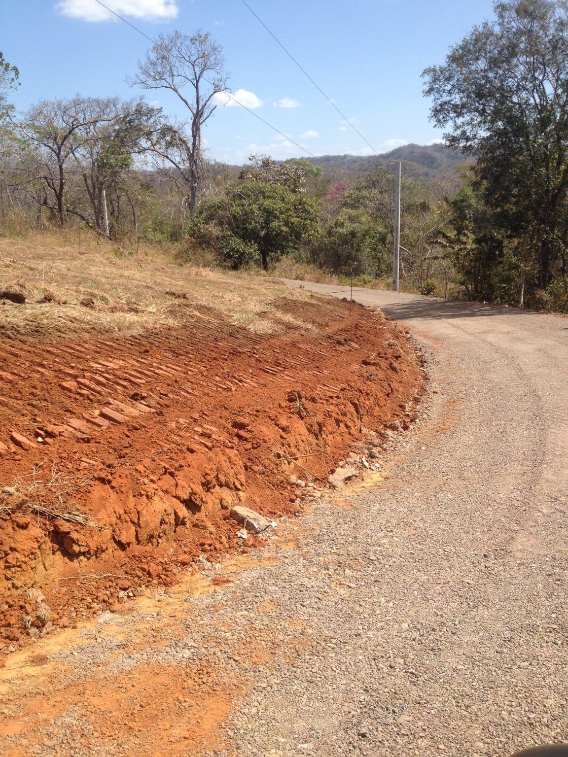 A dirt road curves beside a bank of reddish-brown earth; trees and hills in the background.