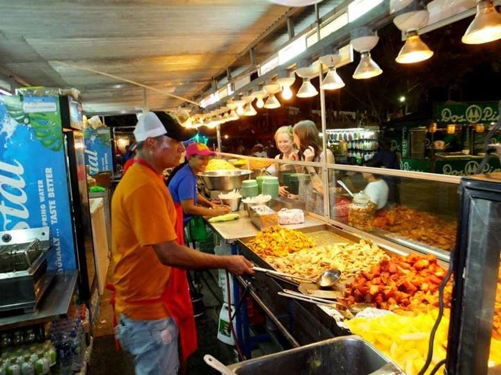 Workers at a food stand prepare and serve dishes to customers. Brightly lit counter displays various foods.