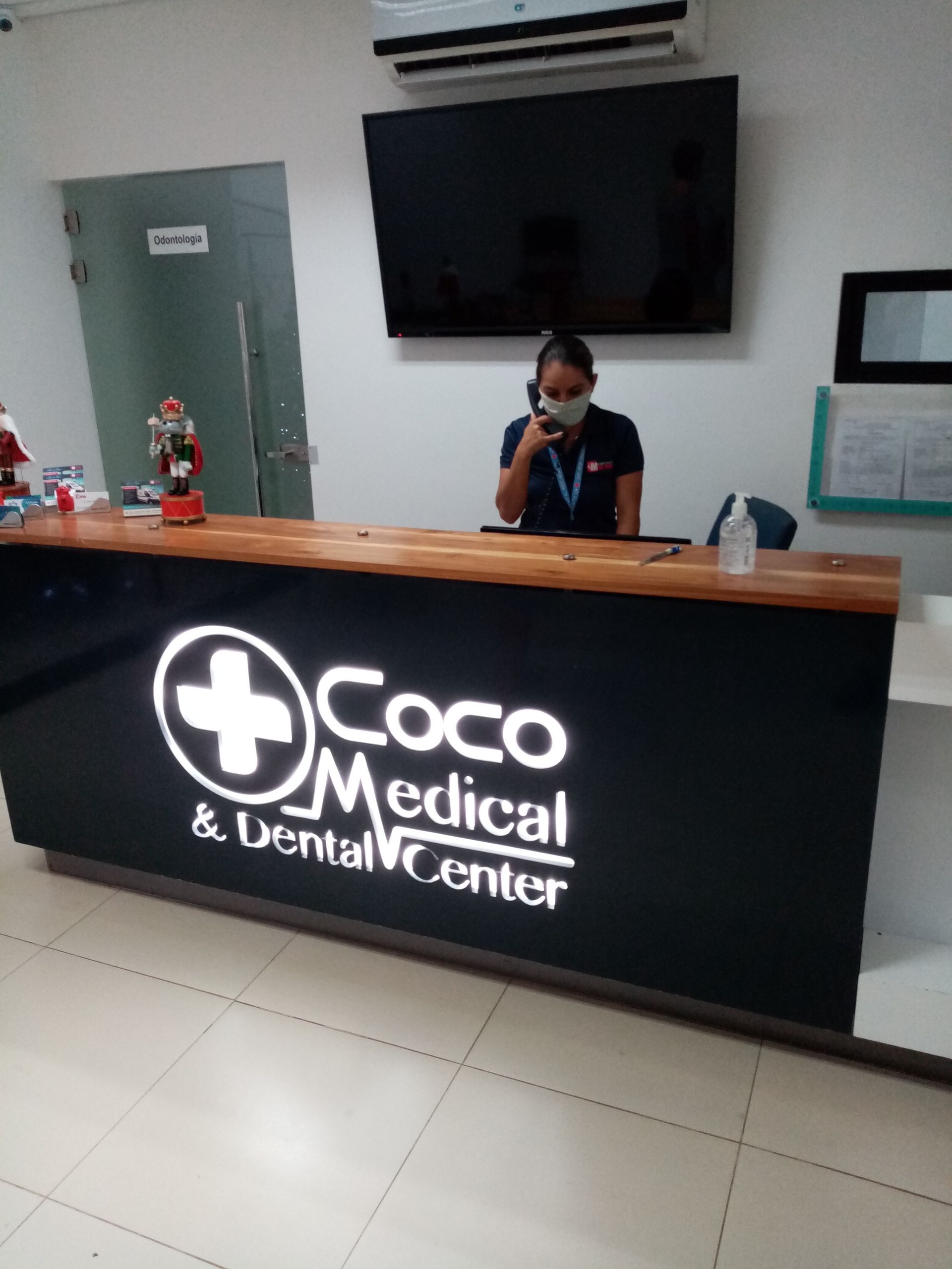 Receptionist at the Coco Medical & Dental Center desk. Woman wearing a mask sits, talking on the phone. Black desk with logo.
