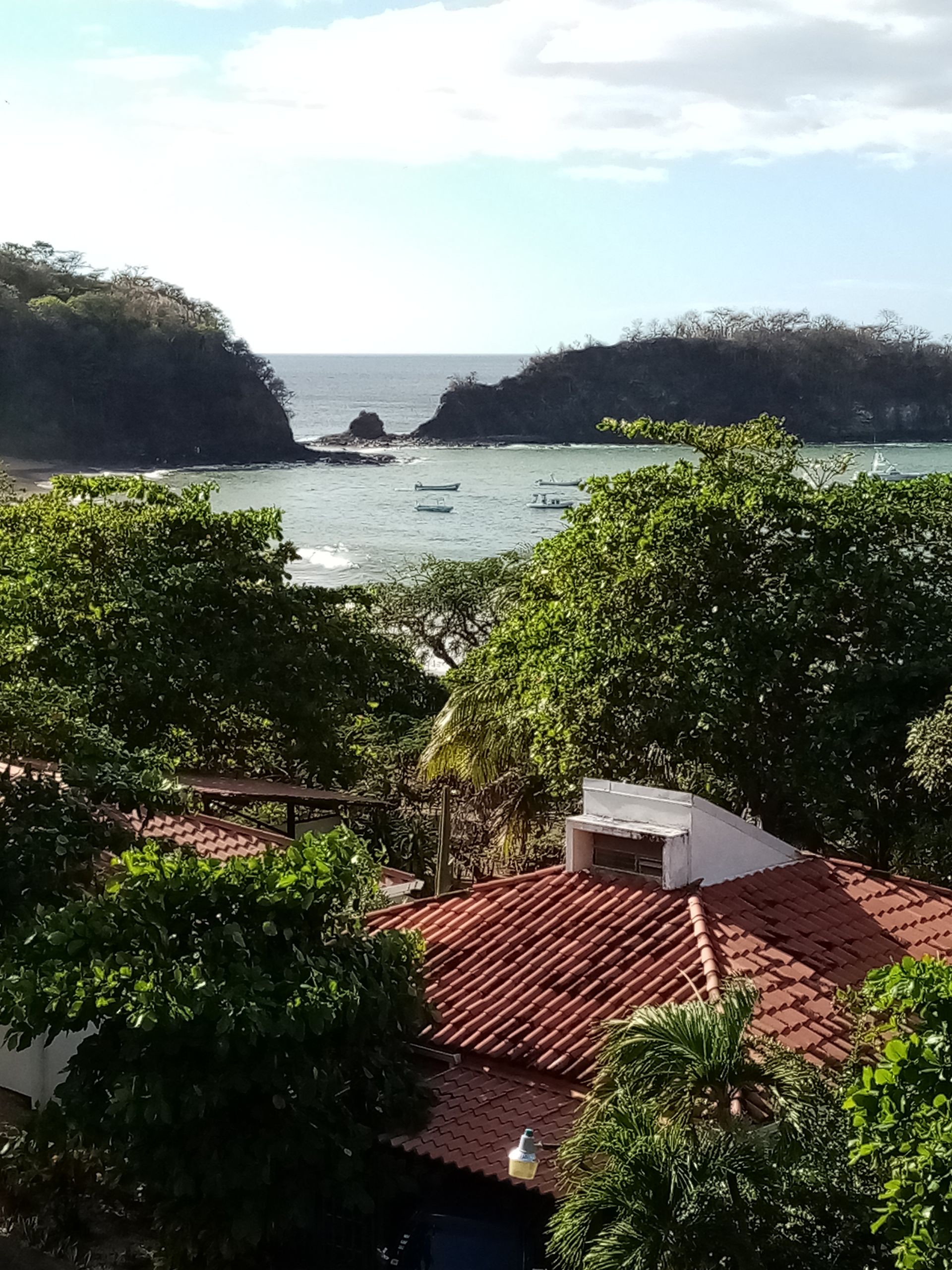 Bay with two rocky islands, visible from a red-tiled roof, framed by green trees. Ocean water and a boat.
