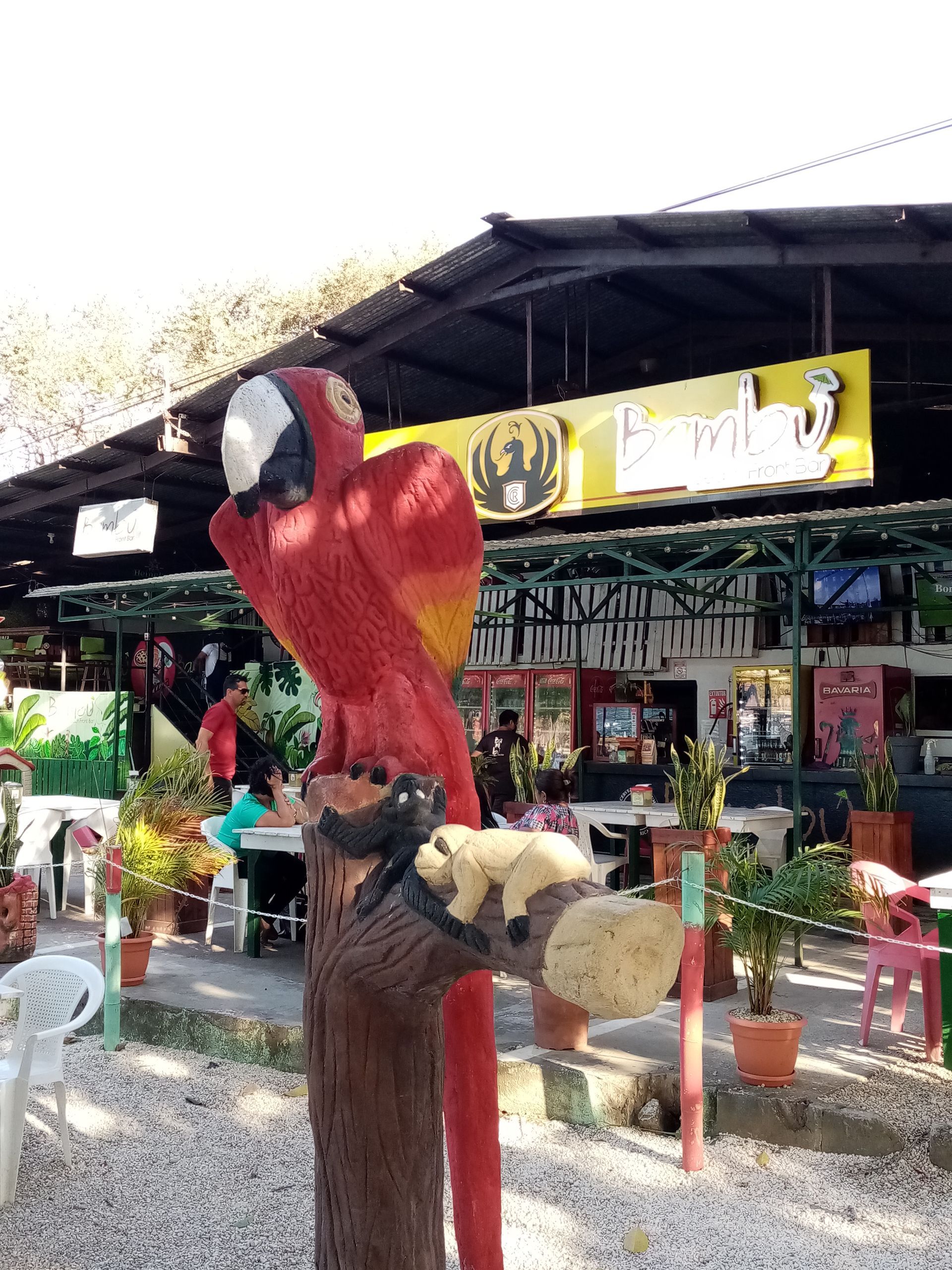 A red parrot statue in front of a restaurant, with people and tables visible.