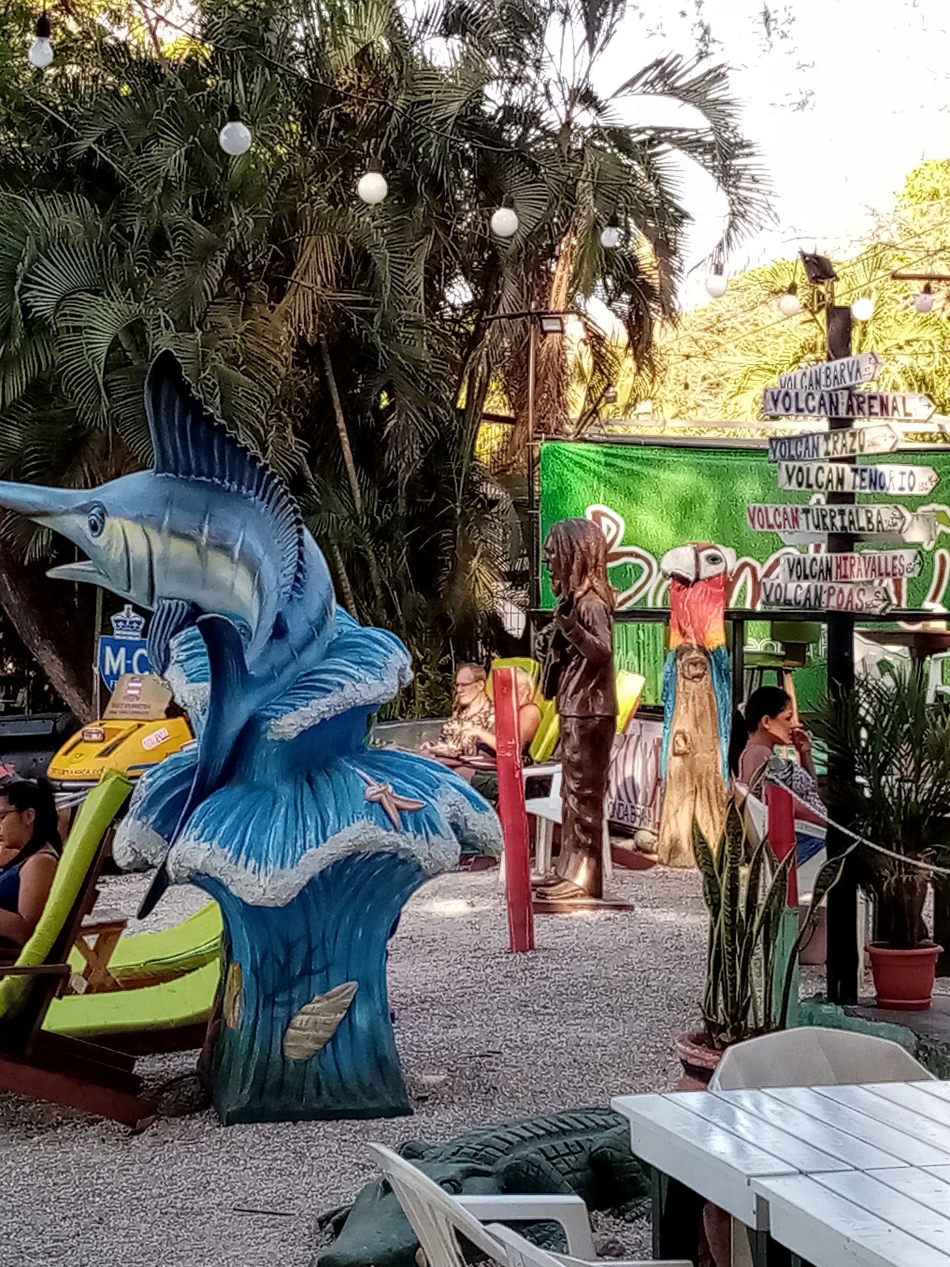 Outdoor area with sculptures, a signpost, and people near a restaurant.