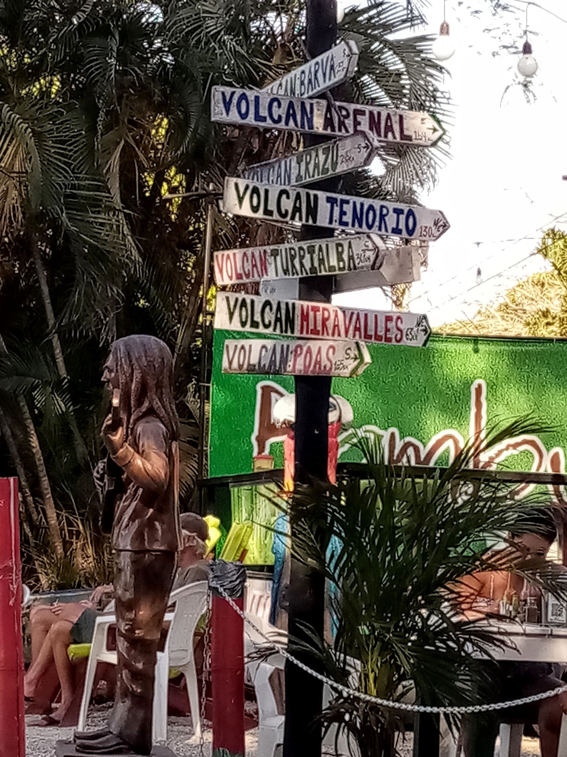 Signpost pointing to various volcanoes, with a statue of a person in front.