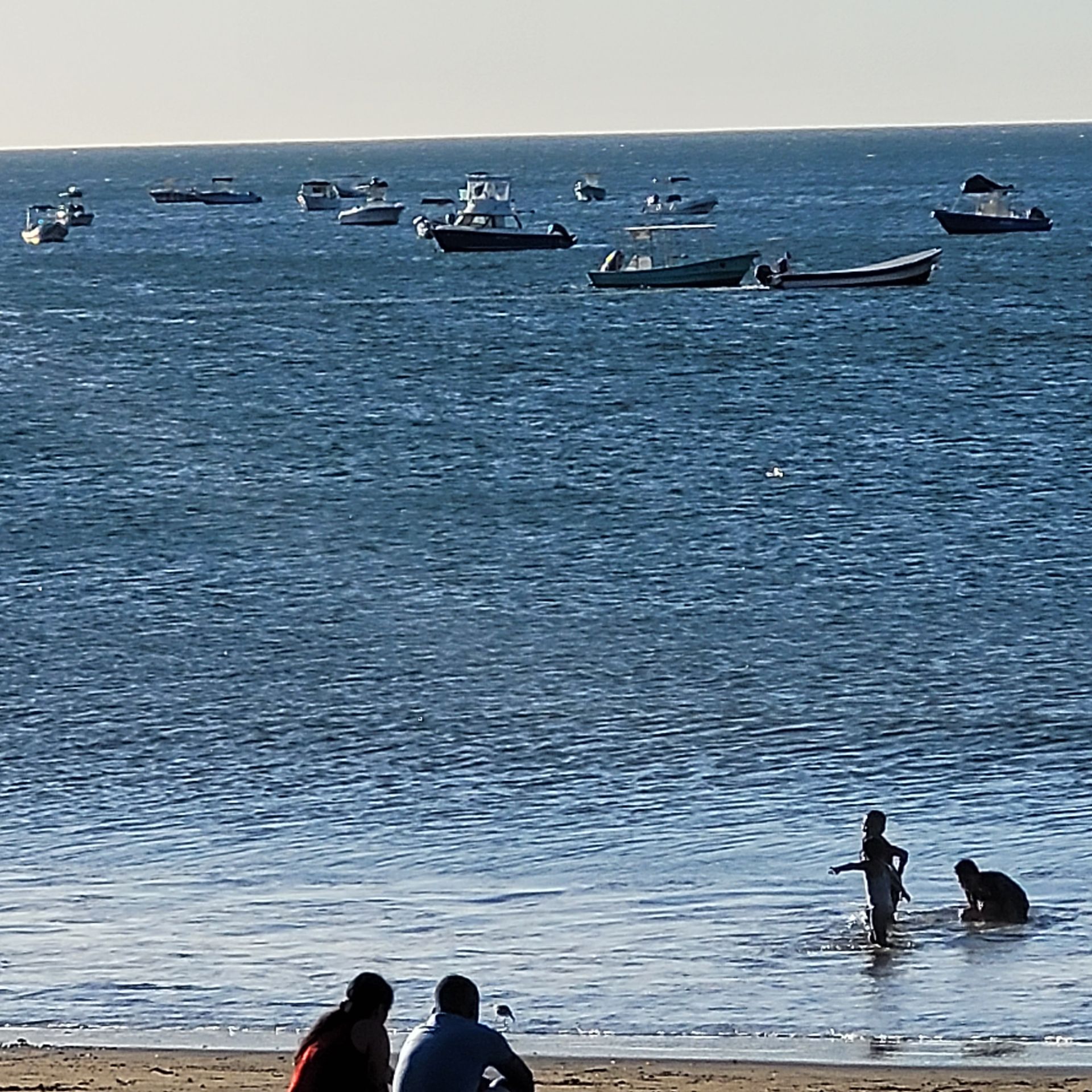 Boats on water; people on beach. Child wading, others sitting. Ocean and sky are visible.