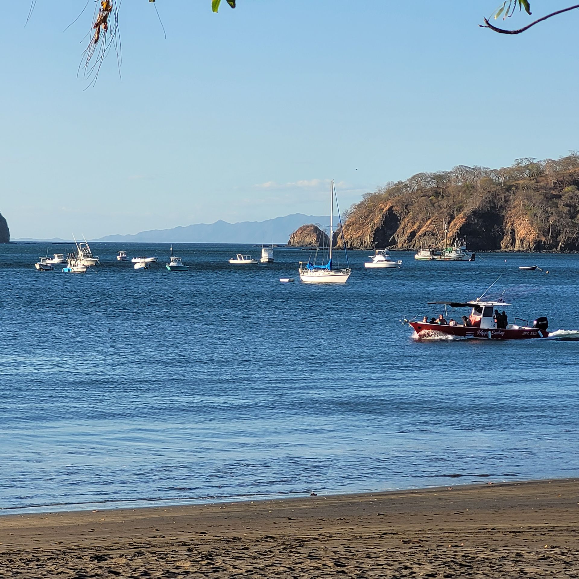 Boats on calm blue water near a beach; a wooded hillside in the distance.