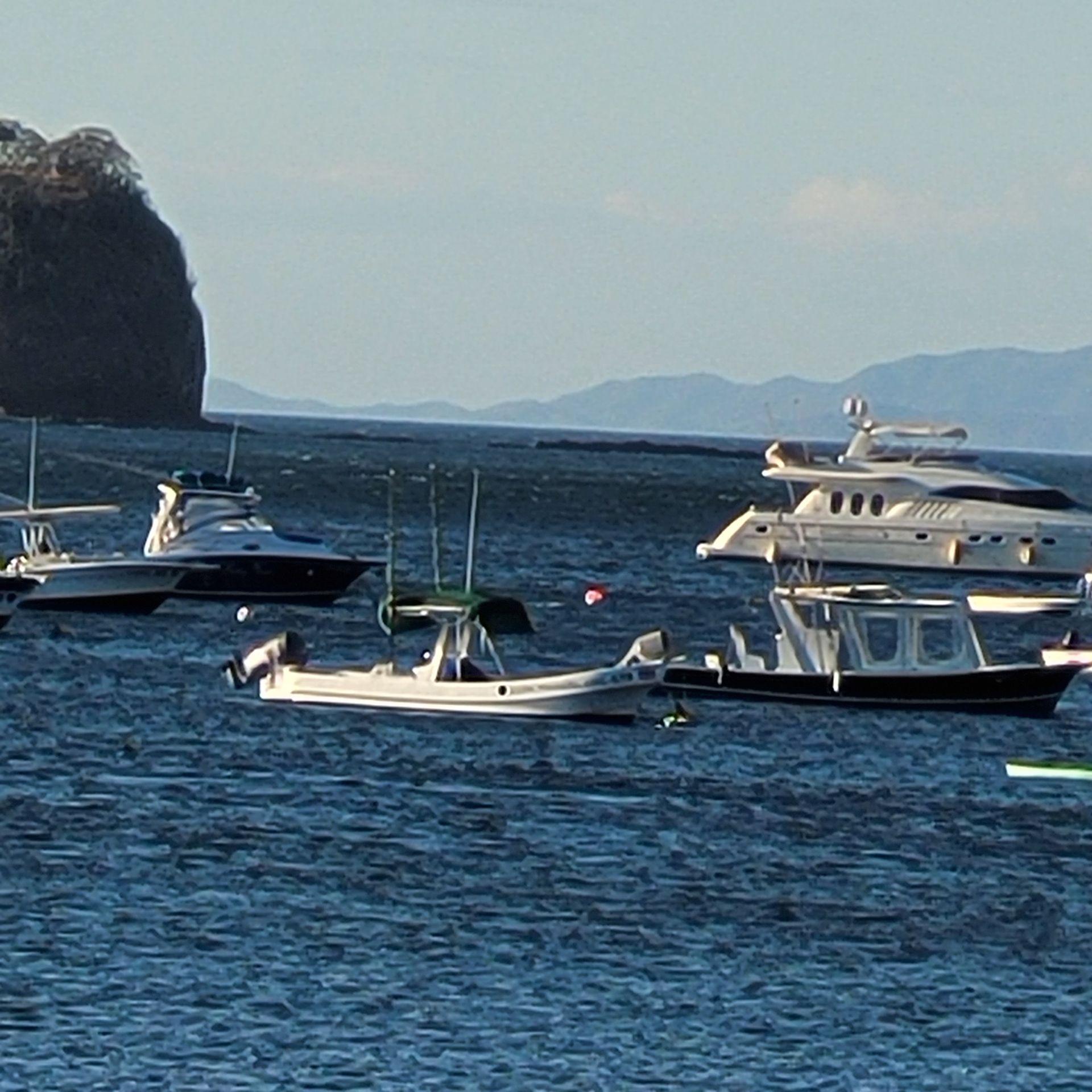 Boats of various sizes docked in a calm bay, with a cliff in the background and mountains on the horizon.