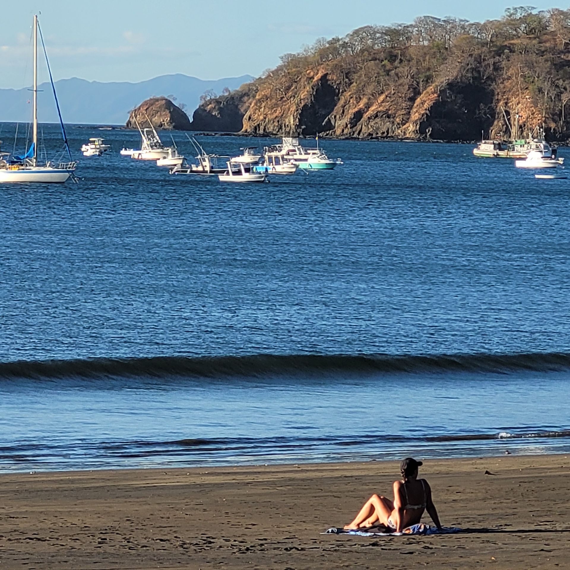 Woman on beach, looking at boats in the bay, brown cliffs in background.