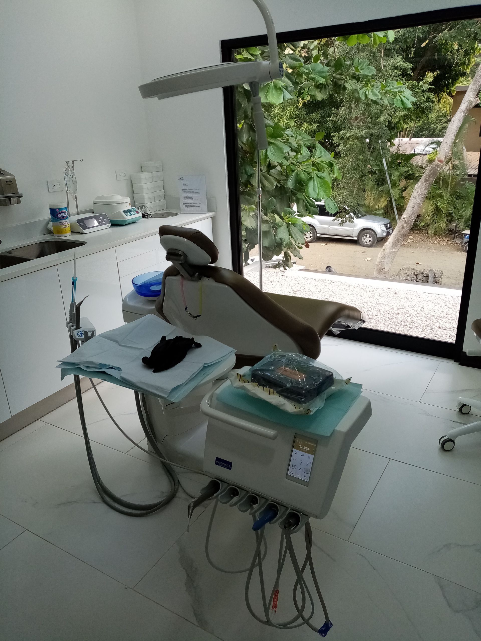 Dental office interior with dental chair, equipment, and window overlooking trees and a street.
