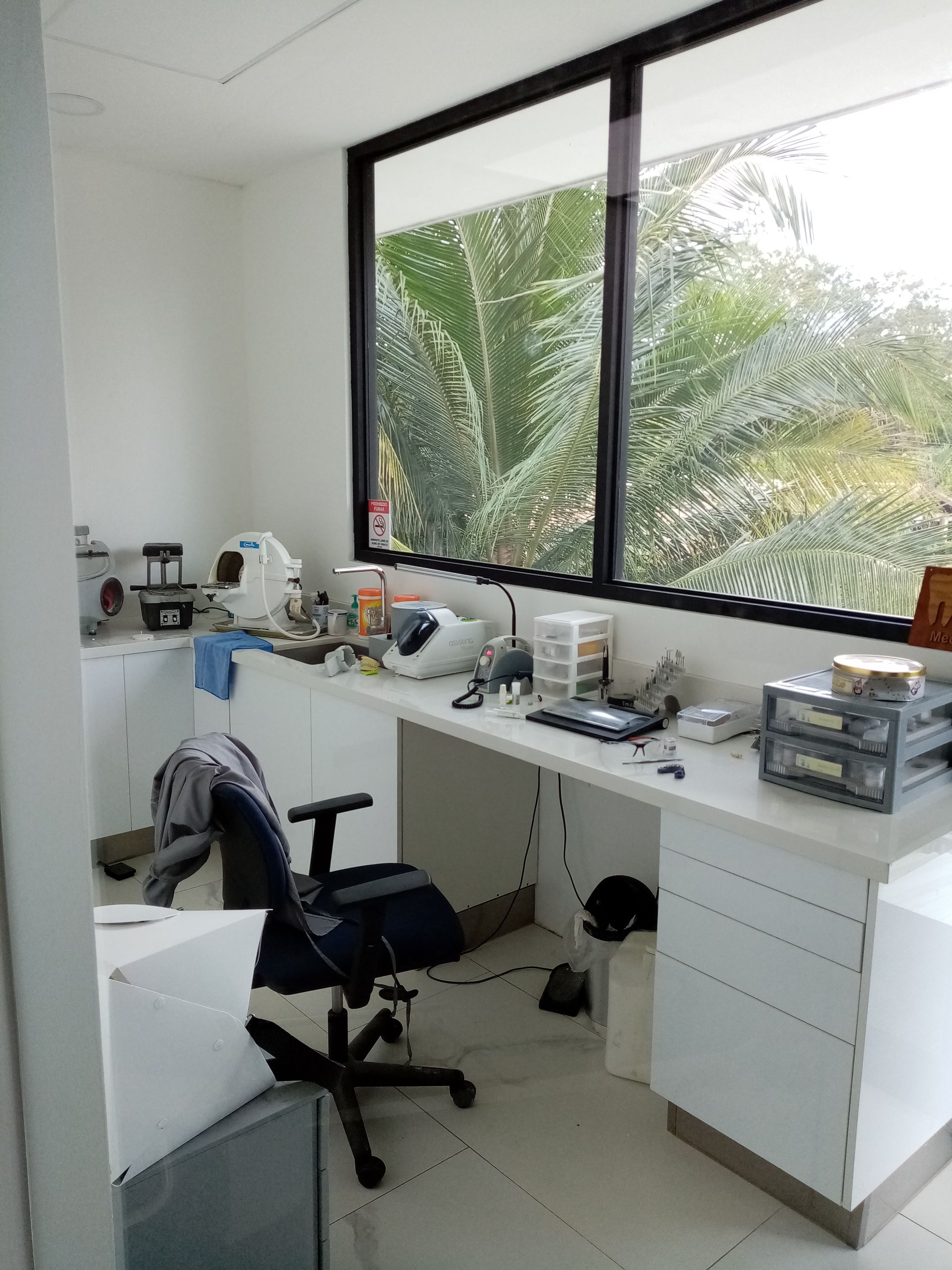 A cluttered white-walled office with large window, desk, chair, and equipment. View of trees outside the window.