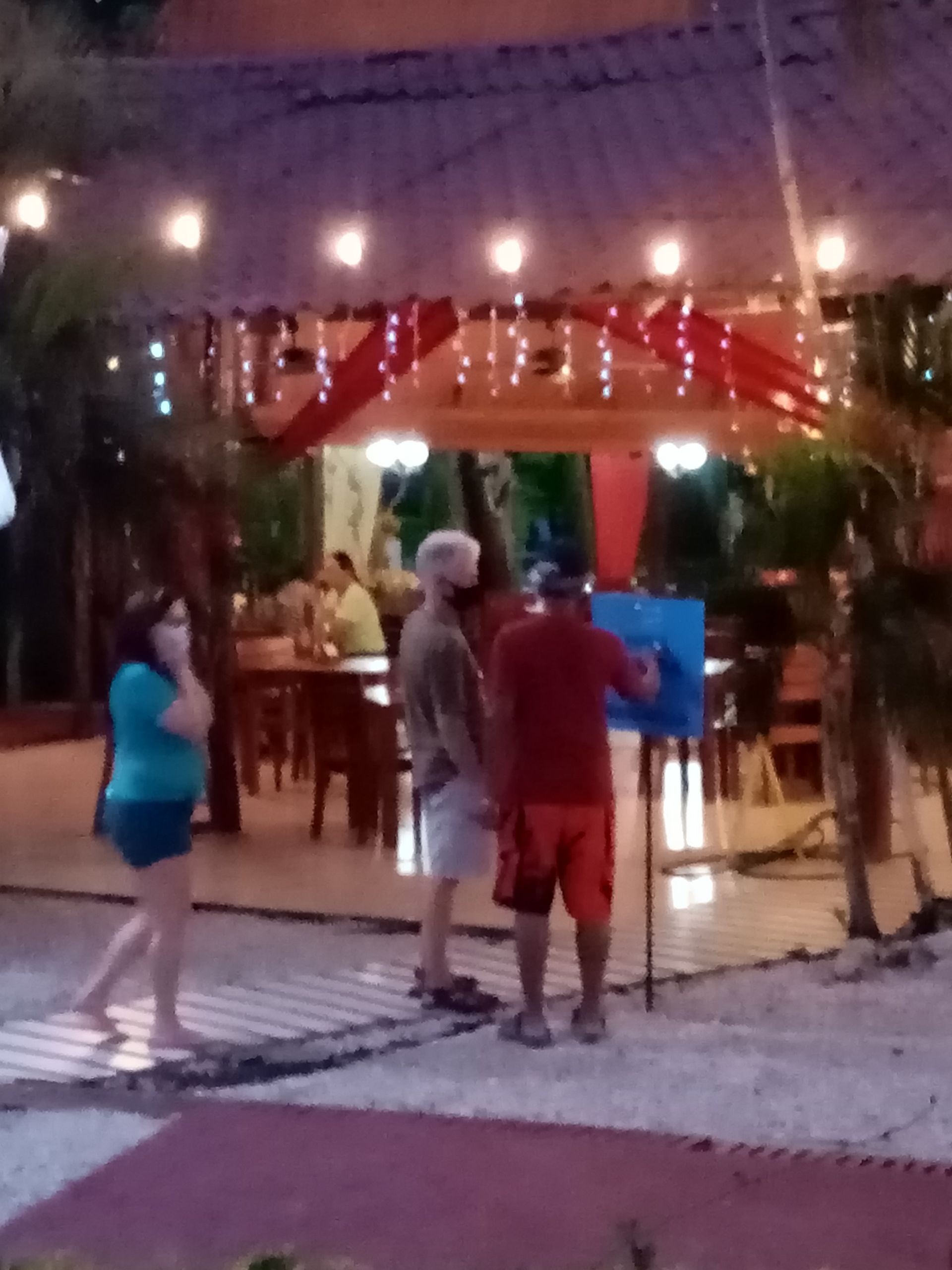 People near a hut decorated with lights; one paints on an easel. Dusk setting.