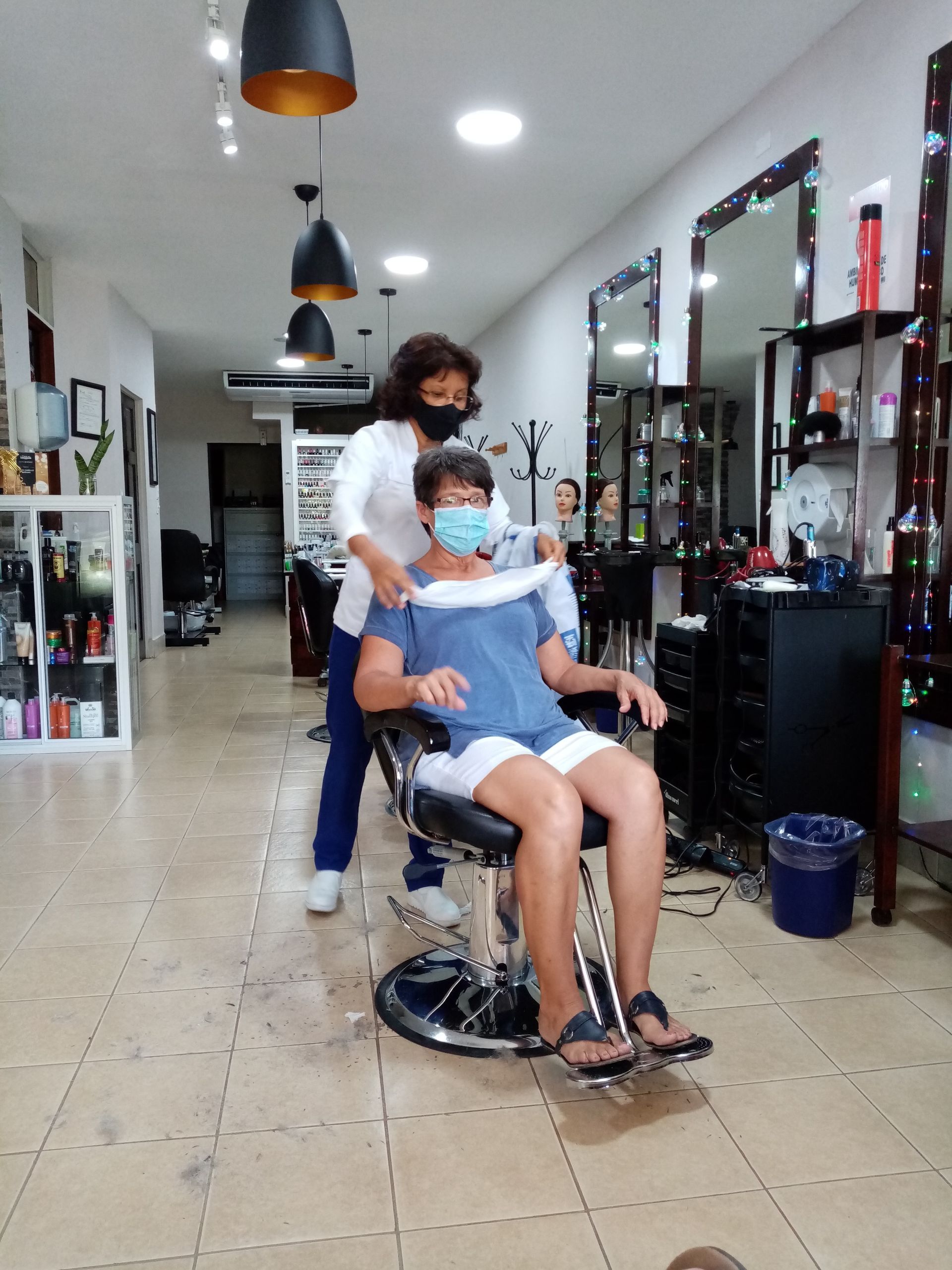 Woman in a salon chair with a stylist putting a neck protector on, both wearing masks.