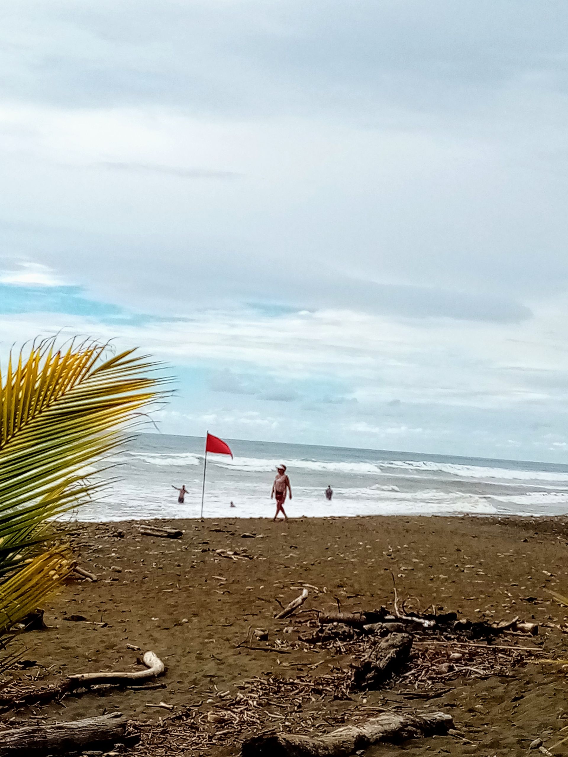 Beach scene with waves, red flag, person walking near shore, cloudy sky.
