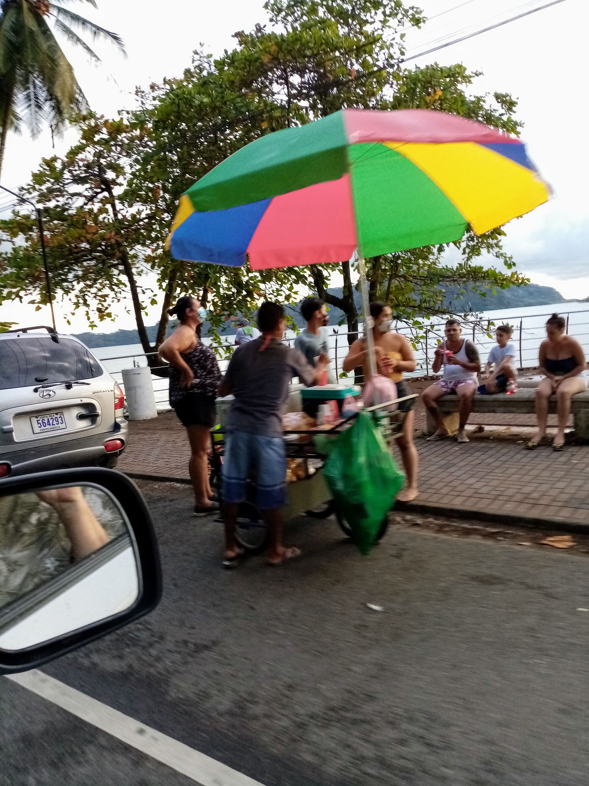 Street vendor with colorful umbrella selling food near water, people gathered nearby.