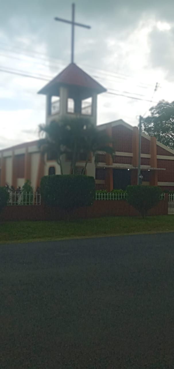 Church with red roof, white steeple, and cross on top. Brick building with green foliage in front.