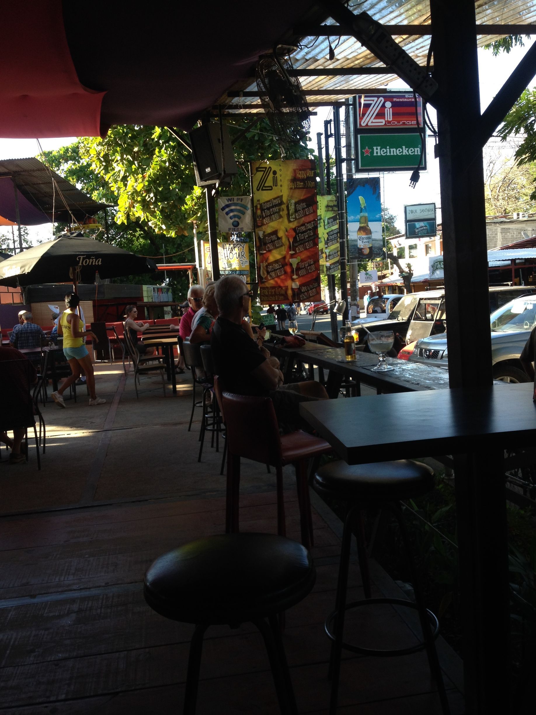 Outdoor bar with stools and tables, people seated. Banners hang from overhead canopy, street visible beyond.