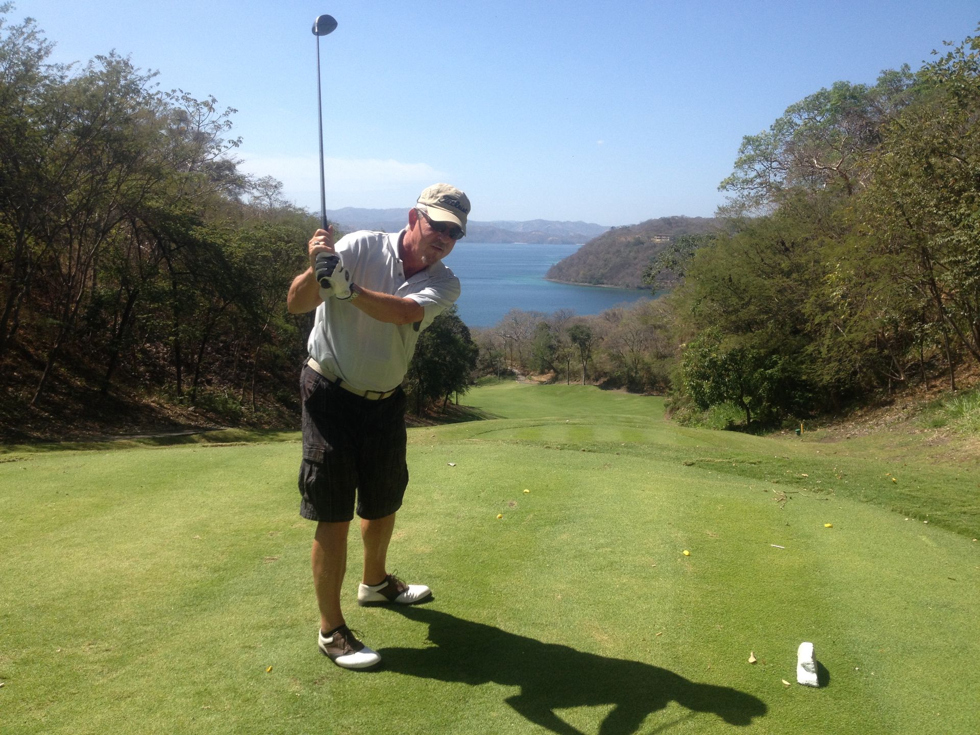 Man swinging golf club on green, overlooking a bay. Bright sunlight.