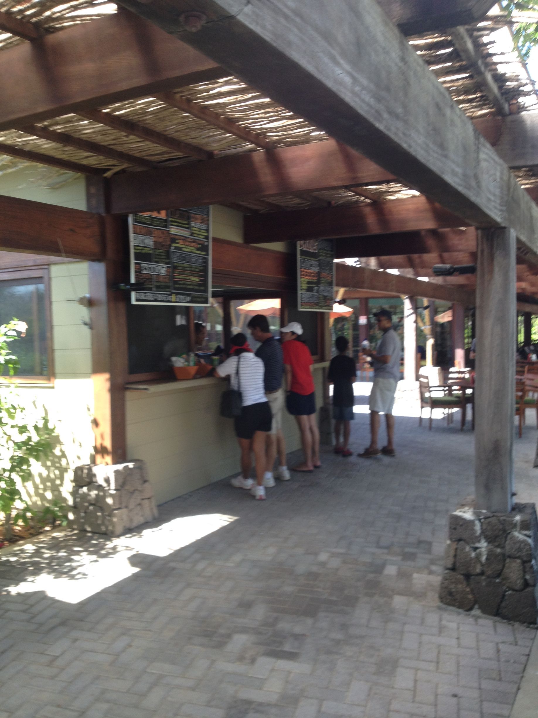 People wait in line at a food kiosk under a shaded pergola on a brick patio.