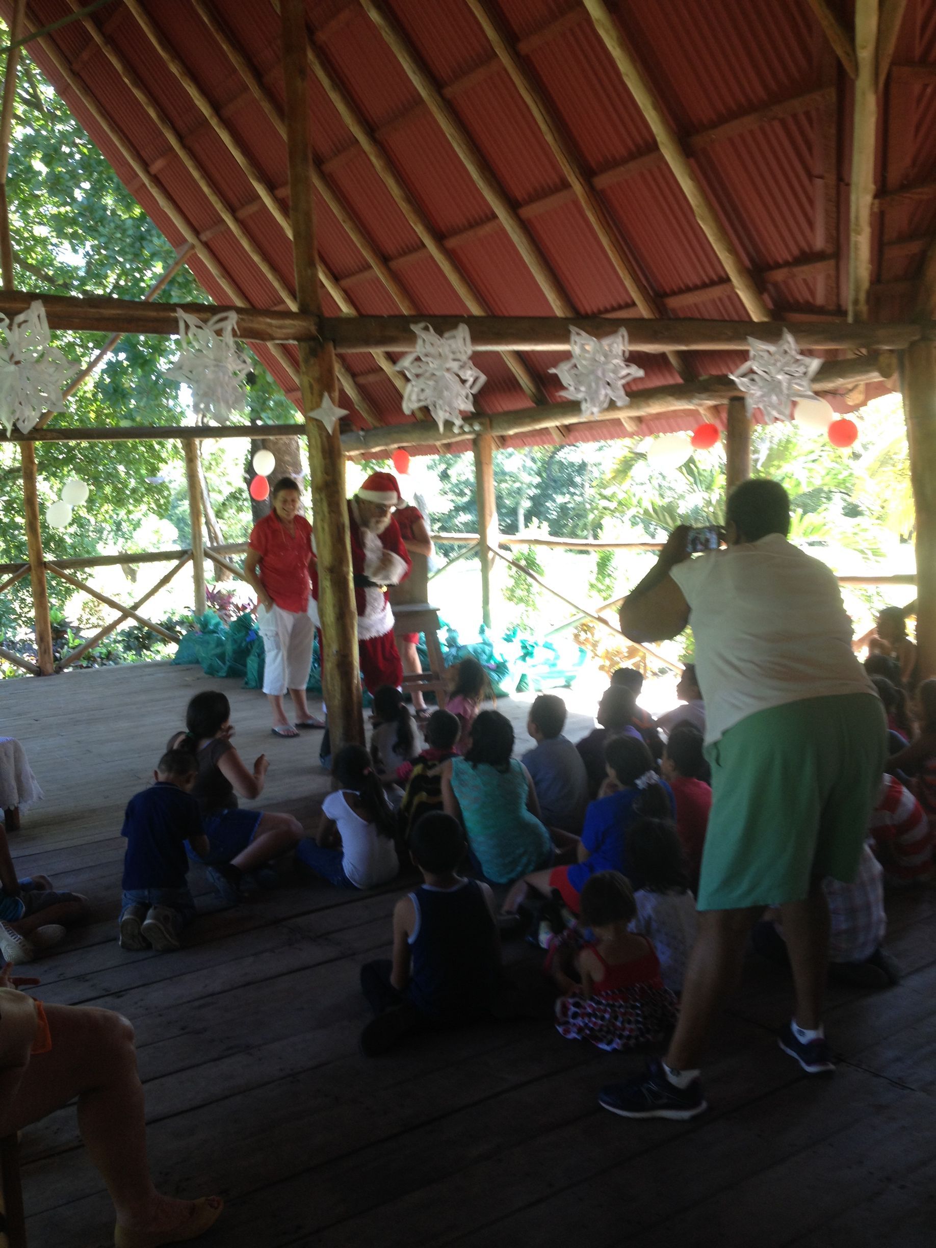 Santa Claus distributes gifts to children gathered under a bamboo and wood canopy, decorated with paper snowflakes.