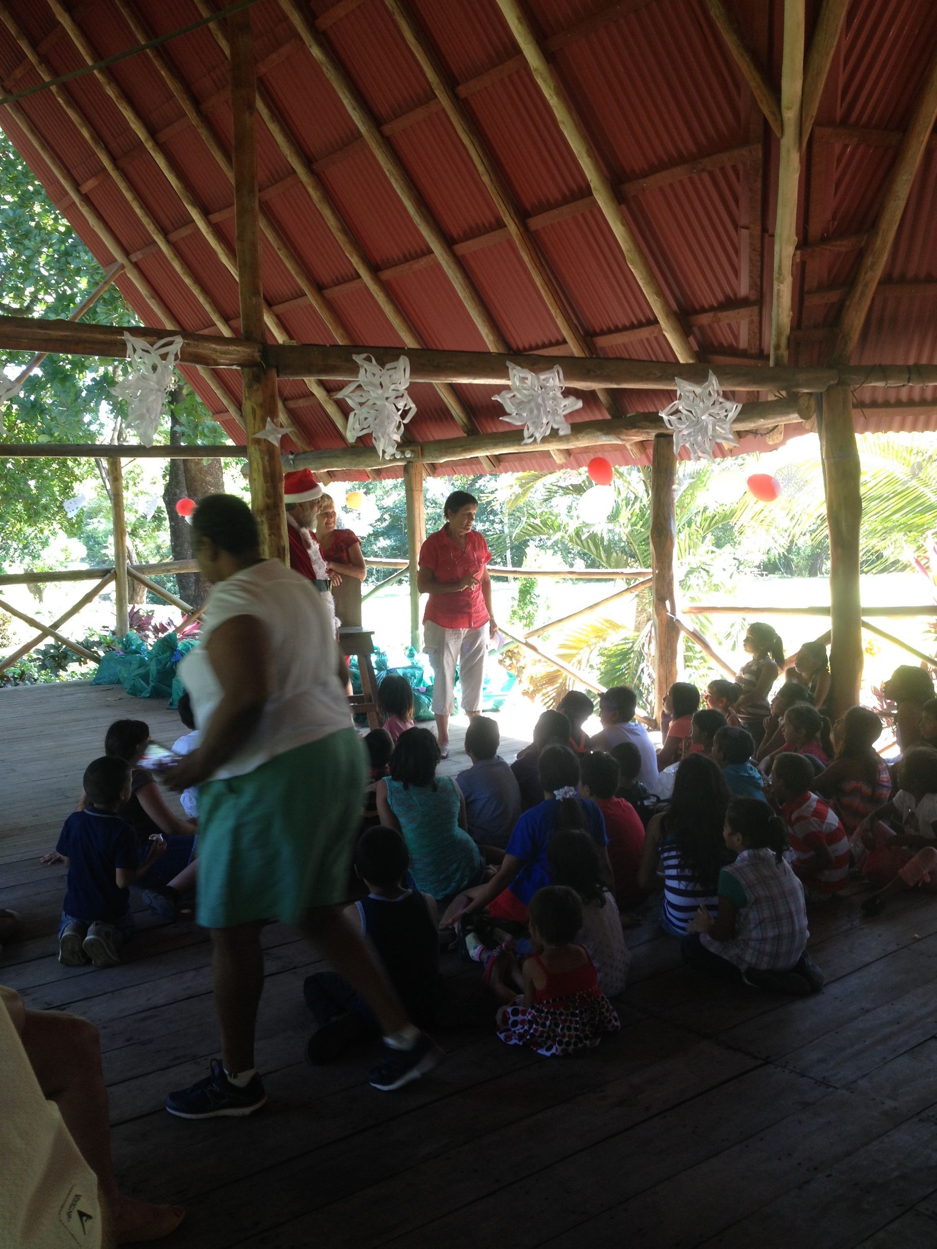 Children seated on a wooden floor, listening to a speaker in an open-air building decorated for an event.