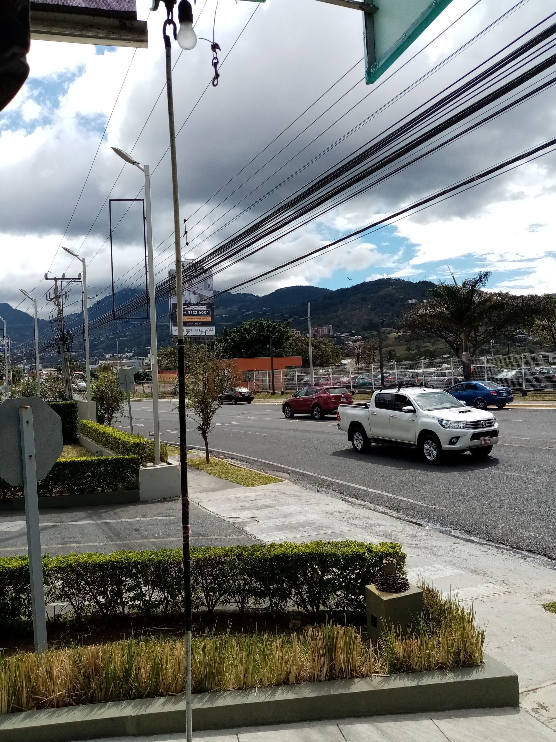 Road with cars, power lines, bushes, sidewalk, and mountains under a cloudy sky.
