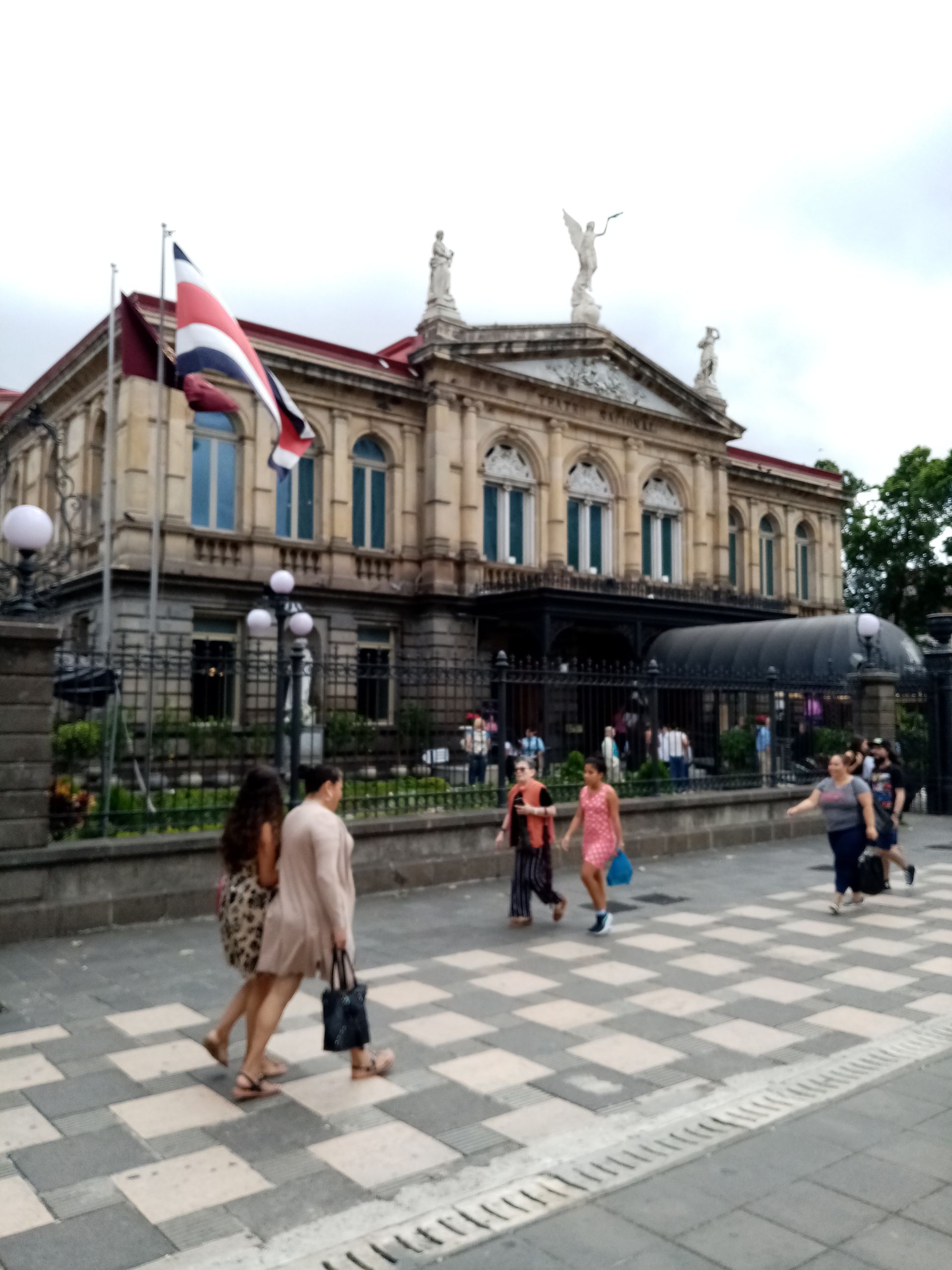 Historic National Theatre of Costa Rica, San José. People walking on a checkered sidewalk. Red, white, and blue flag.