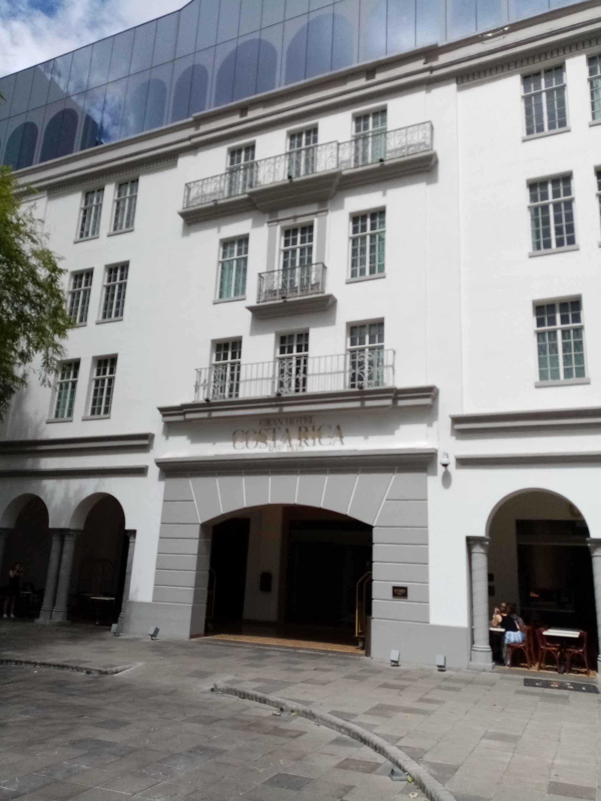 White building with balconies, arched entrance, and glass structure in background.