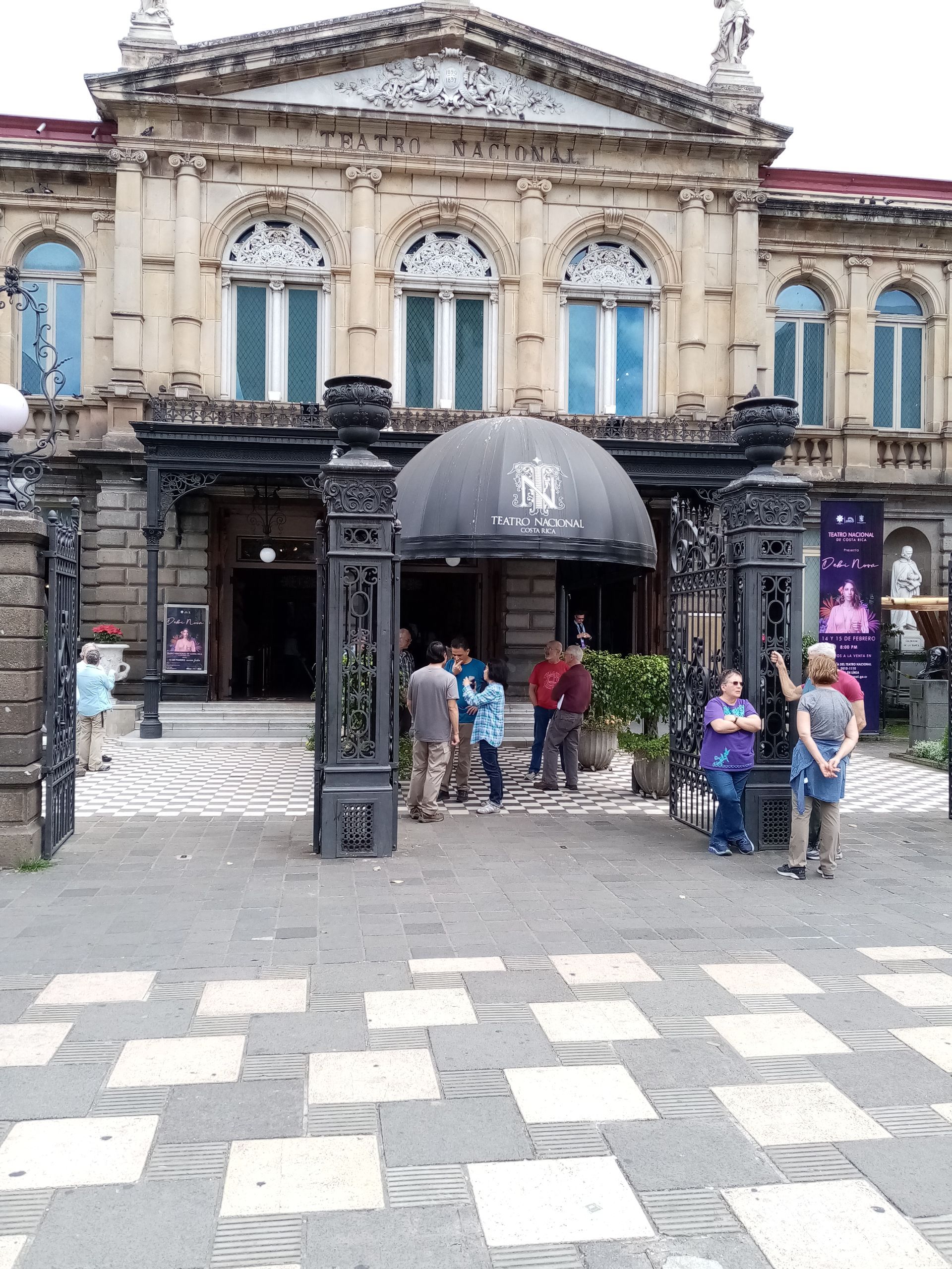 Exterior of a classical building with an ornate entrance, people gathered at the gate.