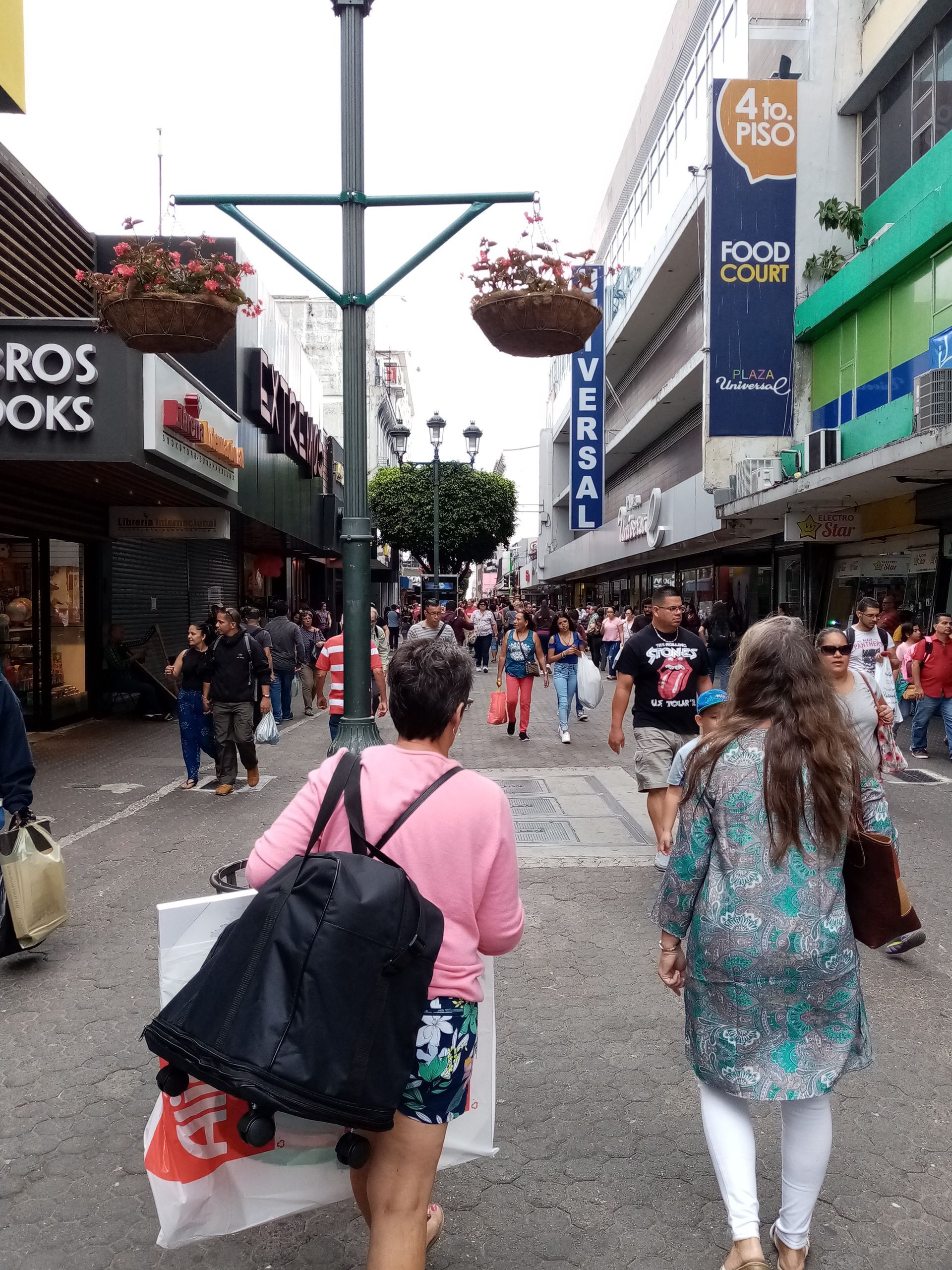 People walking on a pedestrian street lined with shops, hanging flower baskets, and a tall light pole.