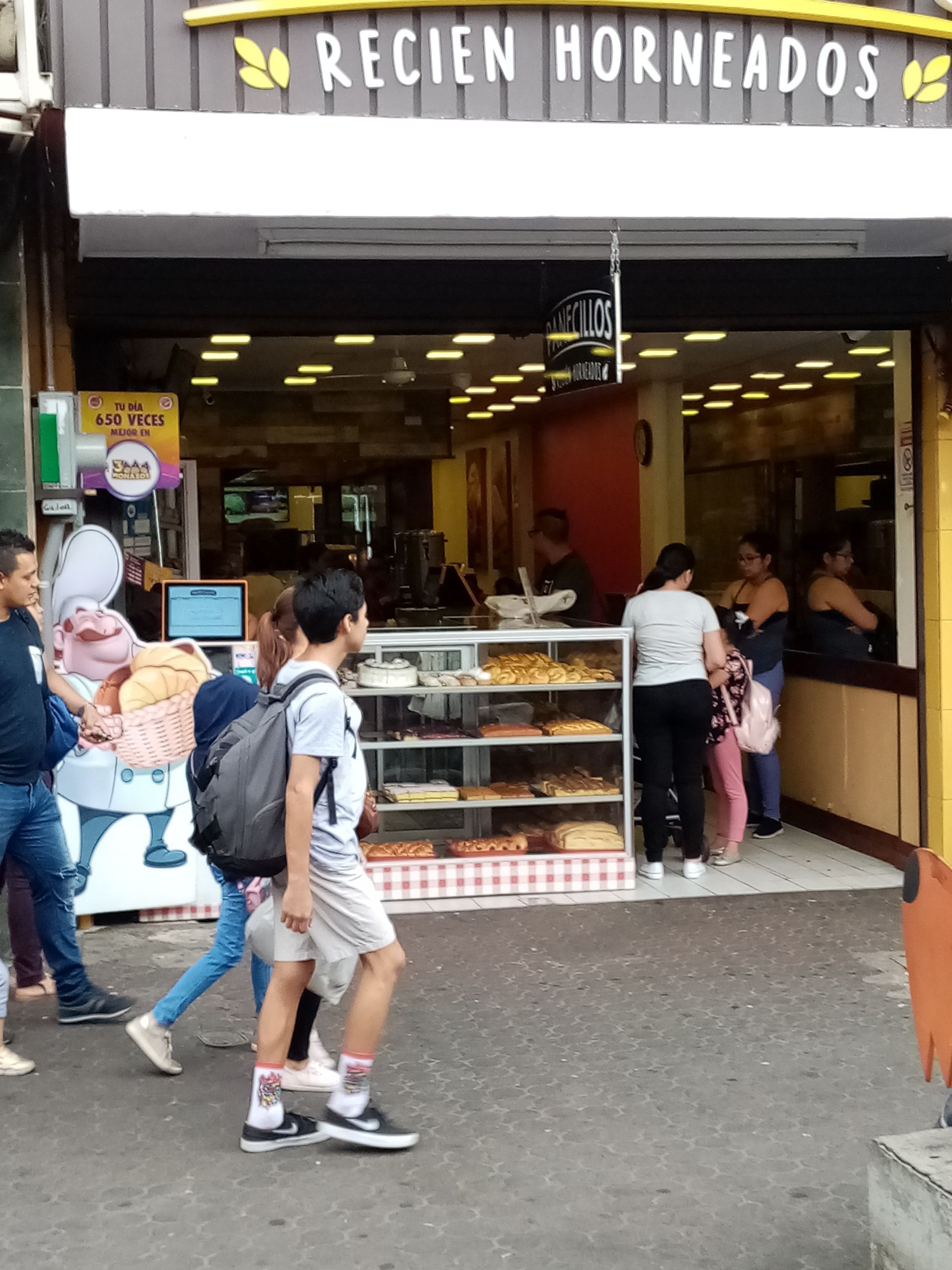 Bakery storefront with people. Display case of baked goods, customers and passersby.