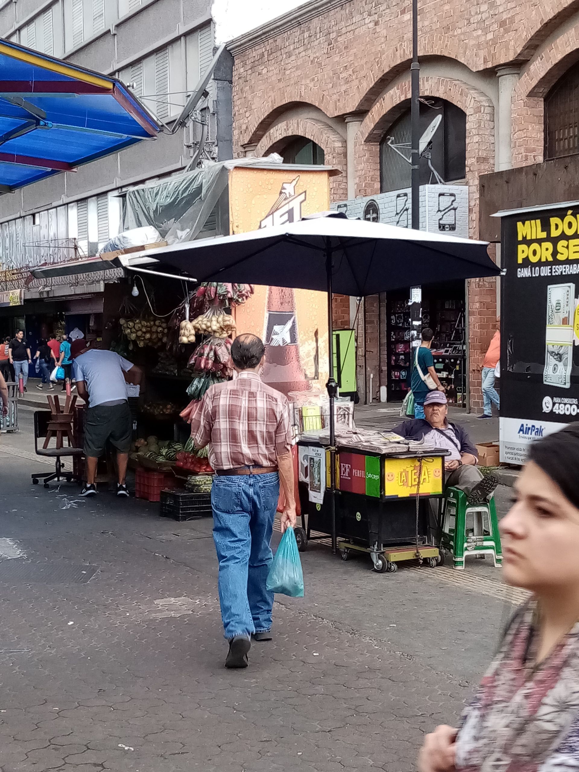 Outdoor market scene; people walking past stalls with goods. Man in plaid shirt walks toward camera.